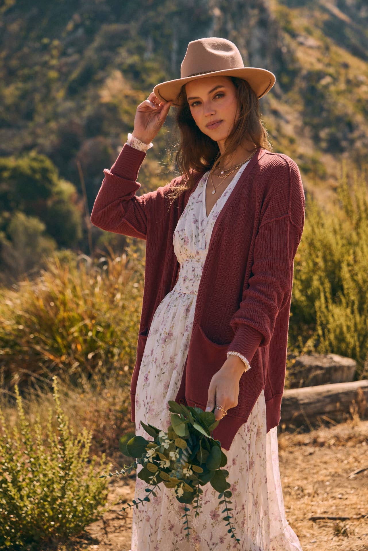 Close-up — mauve knit cardigan layered over cream floral dress with V-neck; tan felt hat; model holding a greenery bouquet outdoors.