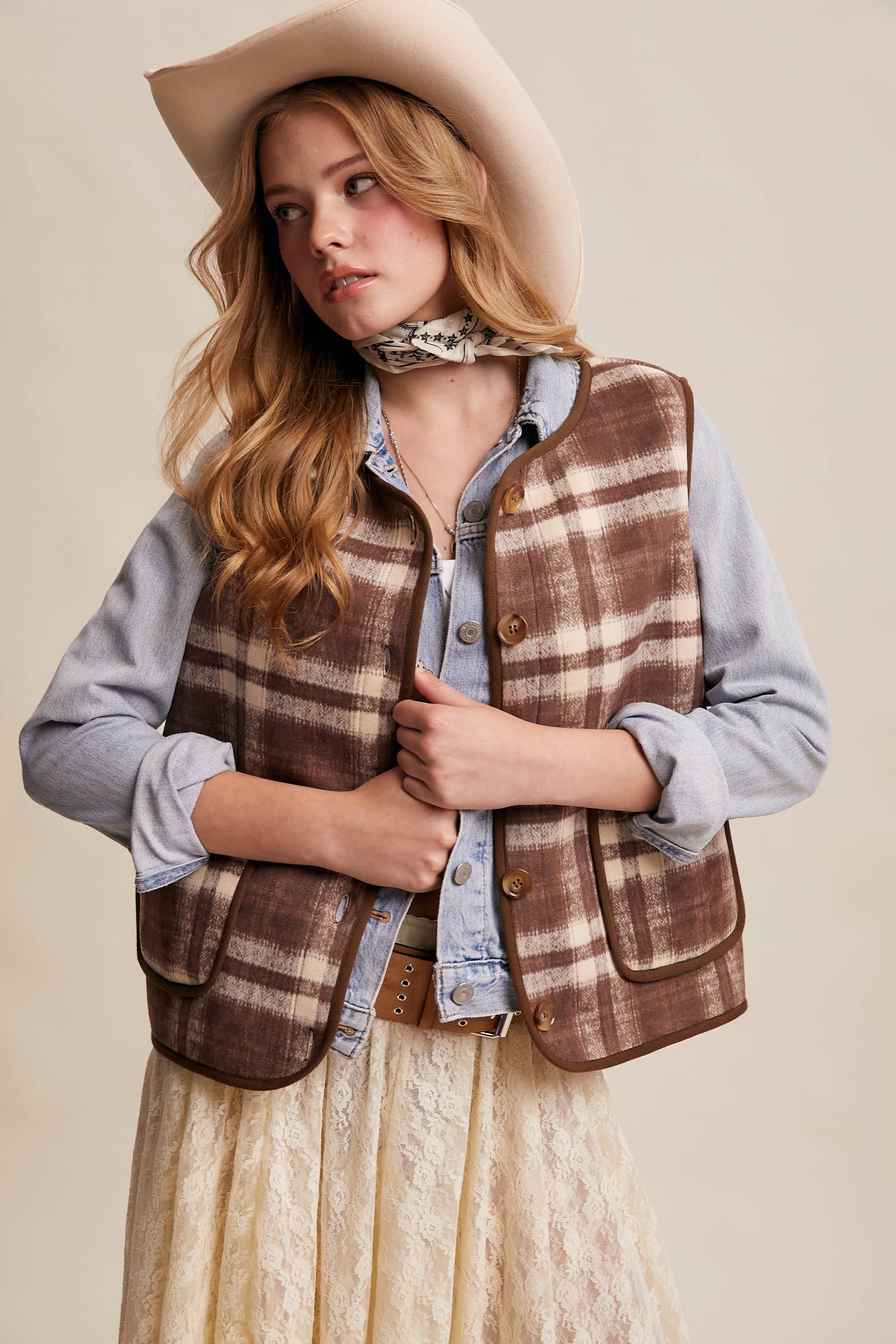 Woman wearing a plaid vest, denim shirt, and beige hat against a beige background