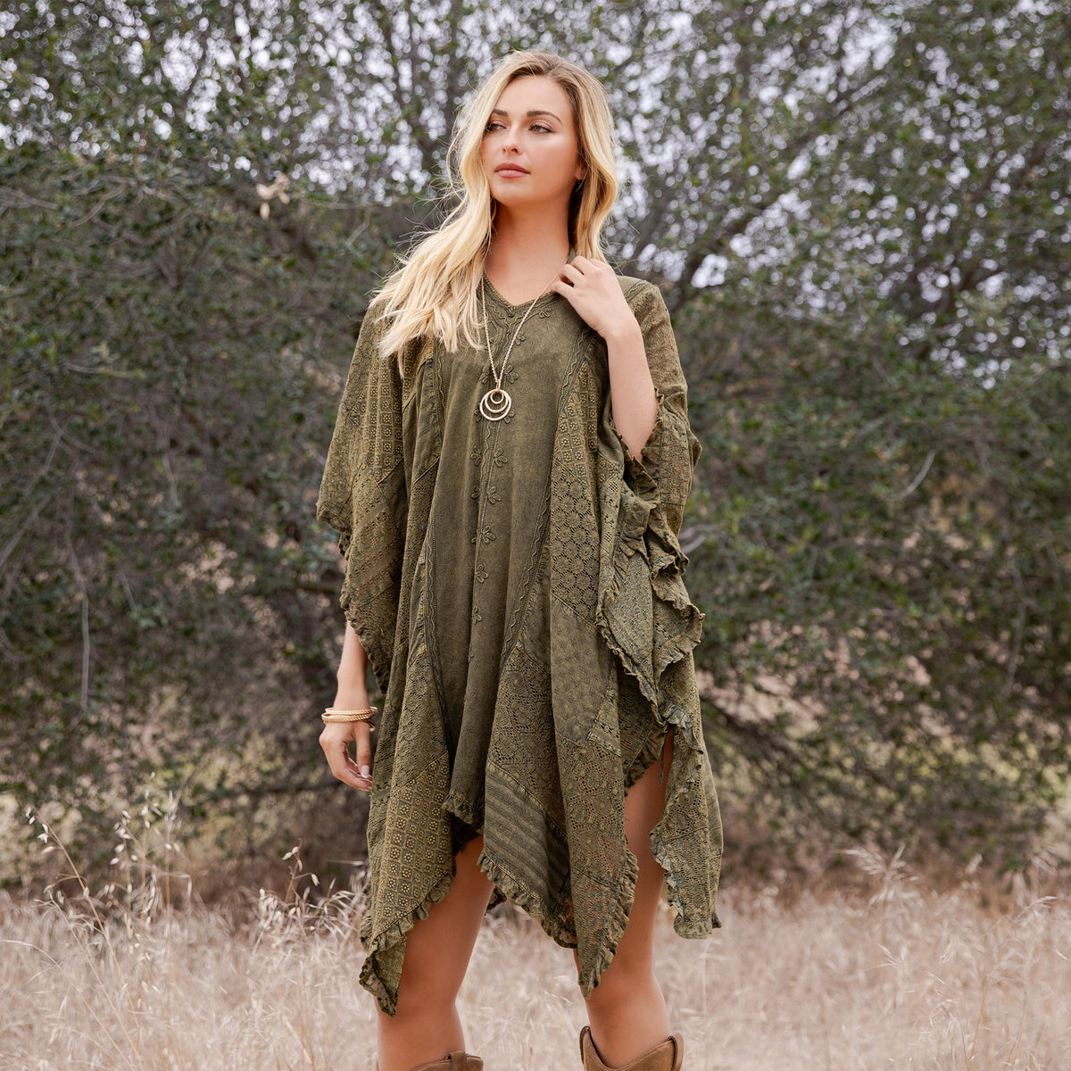 A blonde woman wearing an olive green bohemian-style lace dress with ruffled sleeves, standing in a dry grassy field with trees in the background.