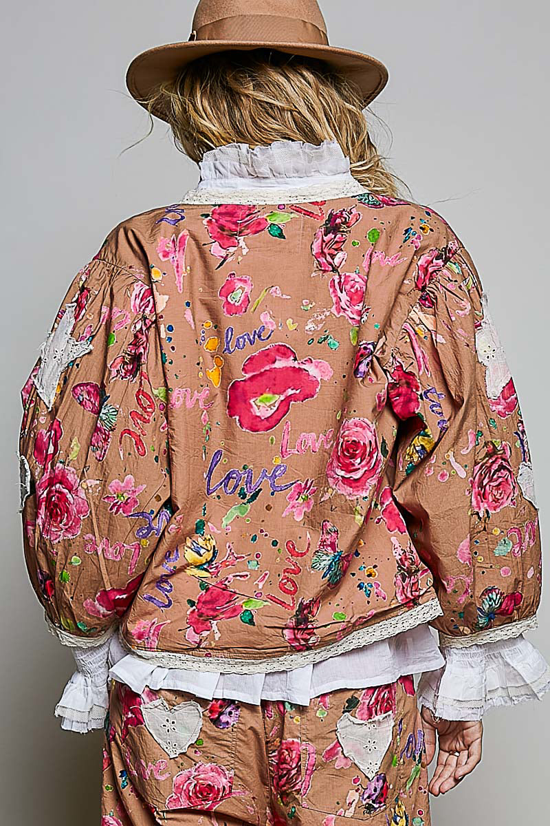 A model shown from the back wearing a brown floral jacket with pink roses, white ruffled trim, and blue 'love' script, paired with matching brown floral pants and a brown hat.