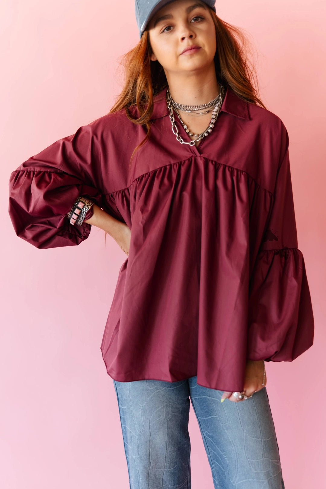 A woman wearing a burgundy long-sleeve blouse with voluminous balloon sleeves and a gathered ruffle detail at the chest, paired with blue jeans and layered silver necklaces, posing against a solid pink background.
