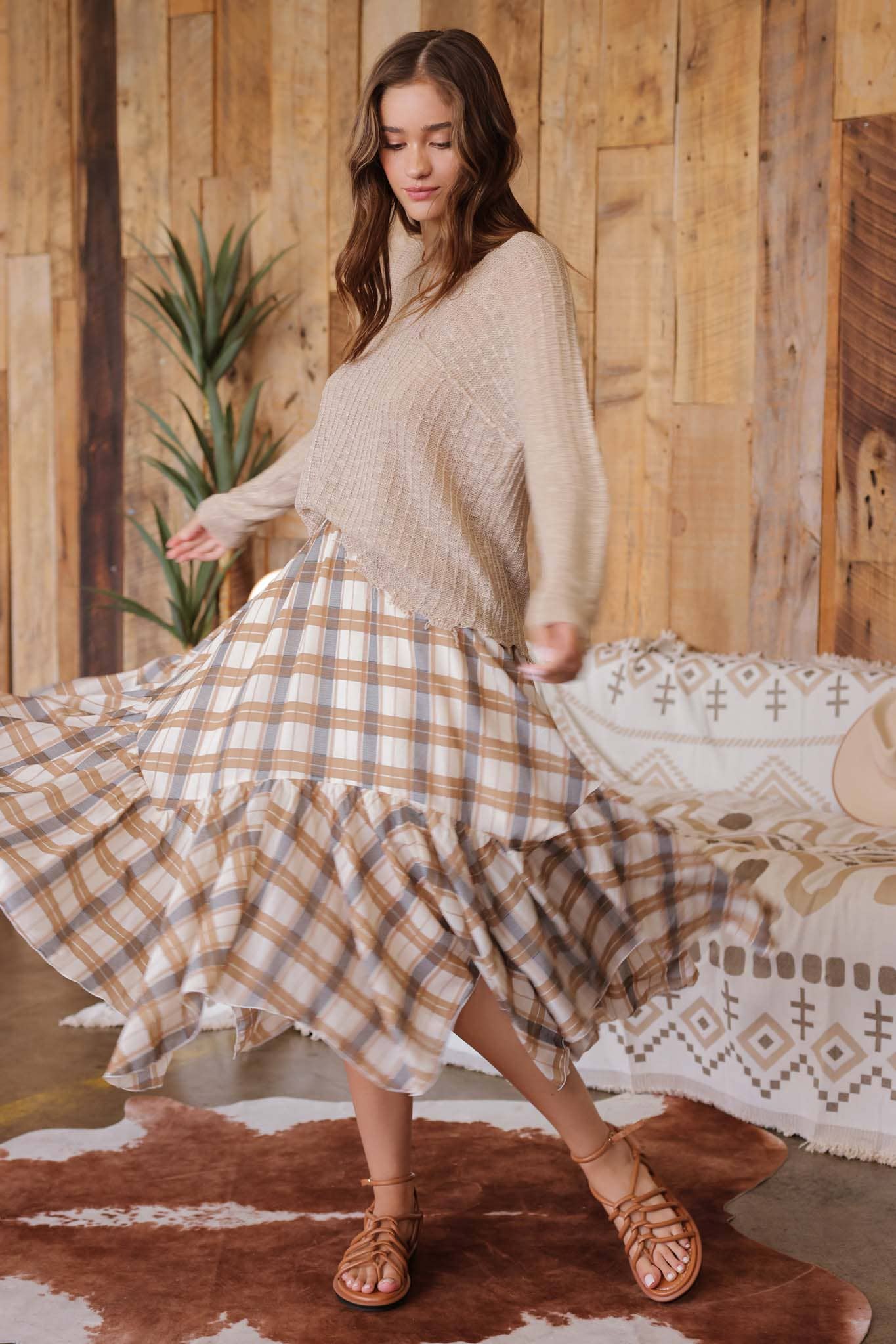 Woman in a beige sweater and plaid skirt standing in a room with wooden walls and a patterned rug.