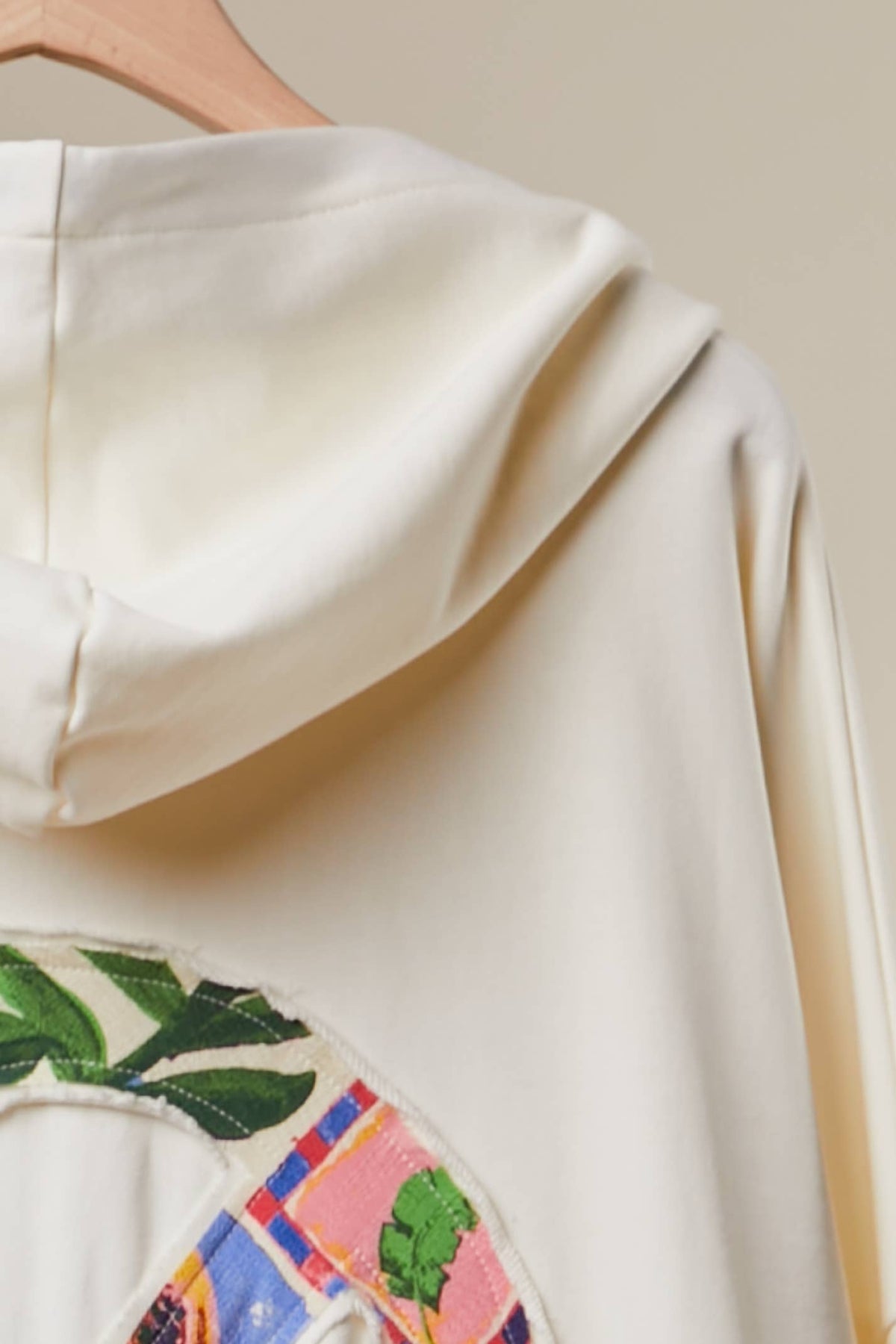 Close-up of a cream-colored hooded sweatshirt hanging on a wooden hanger, featuring a colorful embroidered patch with green leaves and a plaid pattern on the lower left.