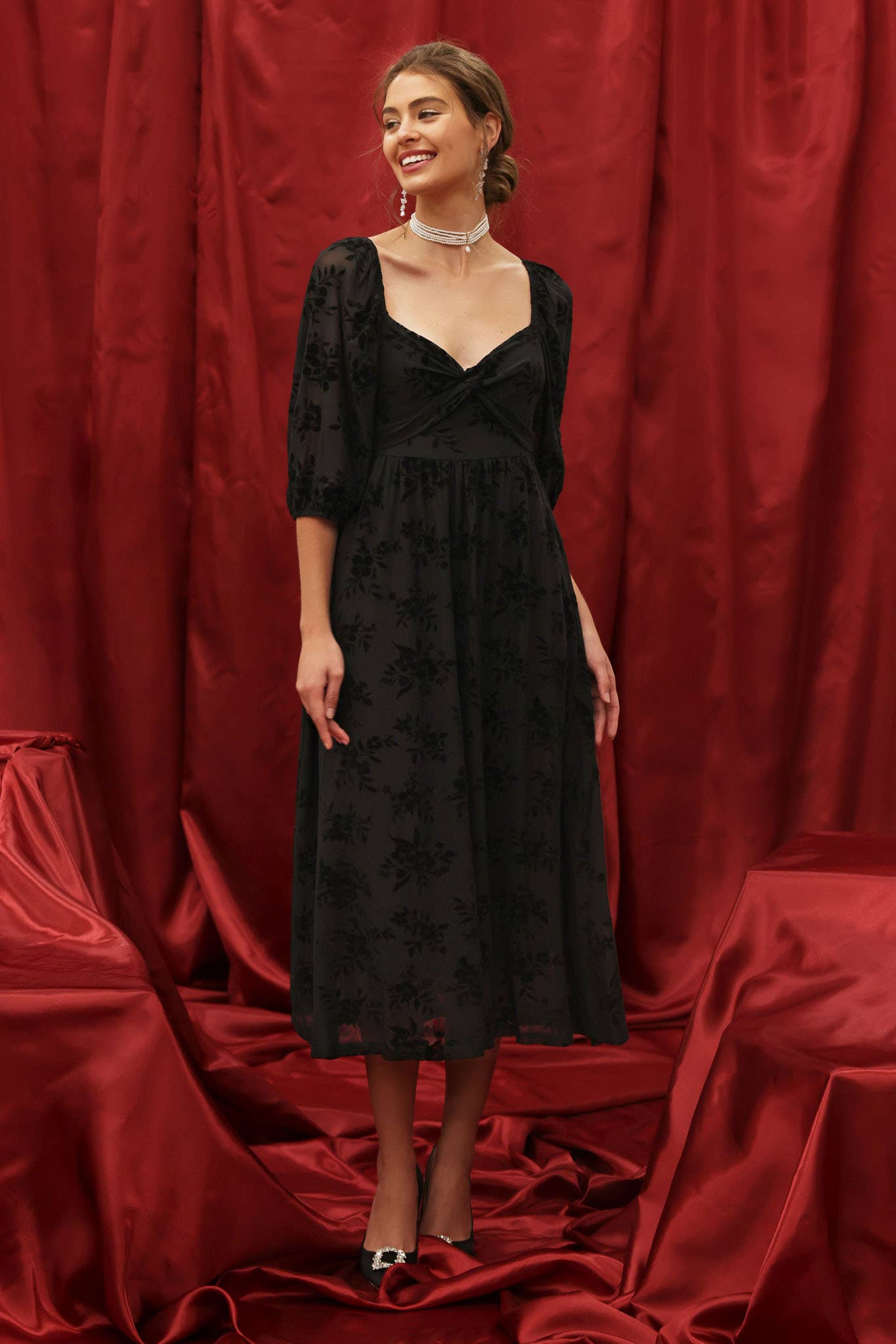 Model wearing a black velvet floral burnout midi dress with sweetheart neckline, twist-front bodice, and 3/4 puff sleeves with elastic cuffs, styled for an elegant holiday look against a red backdrop.