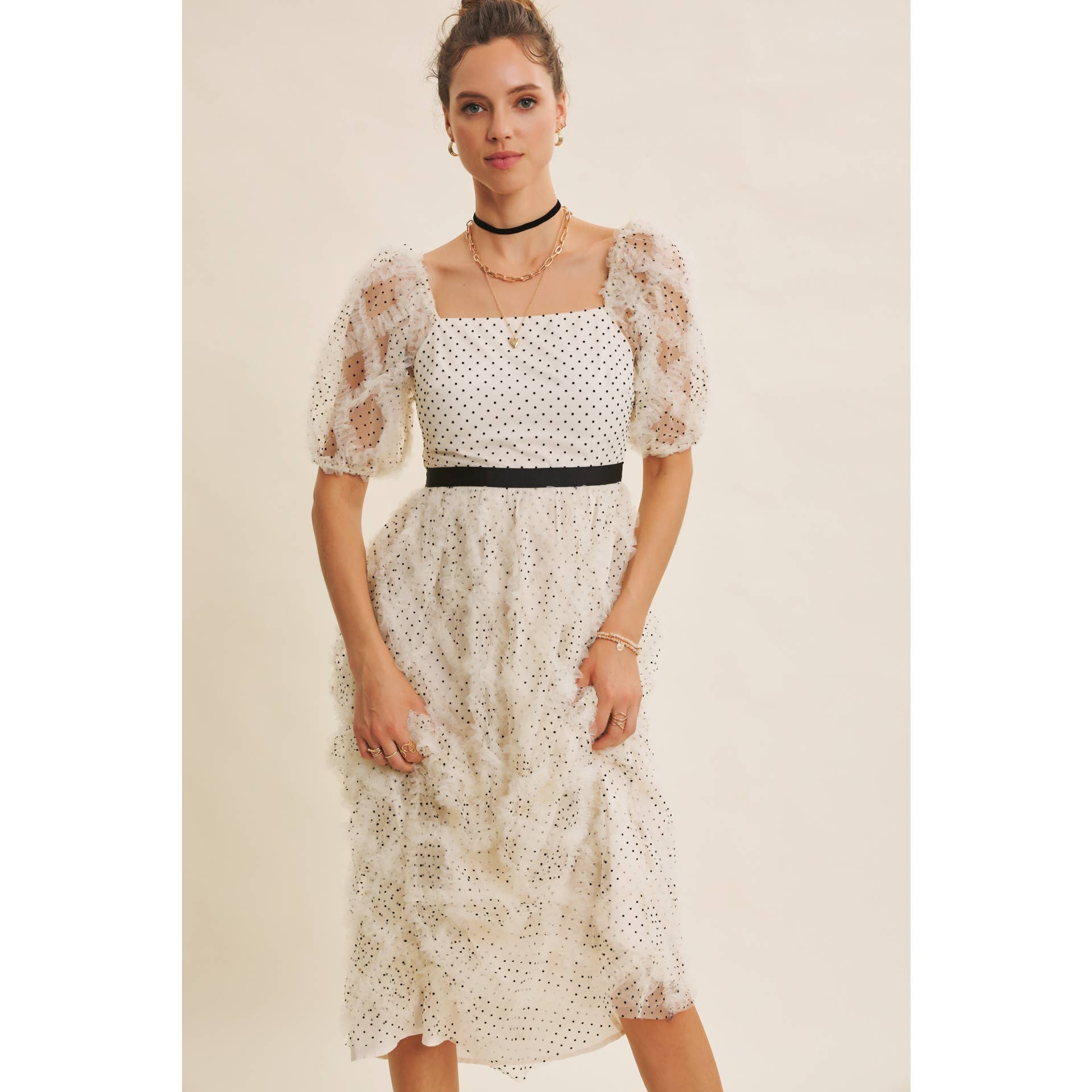 Front view — cream polka-dot tulle midi dress with puff sleeves and black belt; styled with black hat, ankle boots, pearl clutch; leaning by wooden doors.