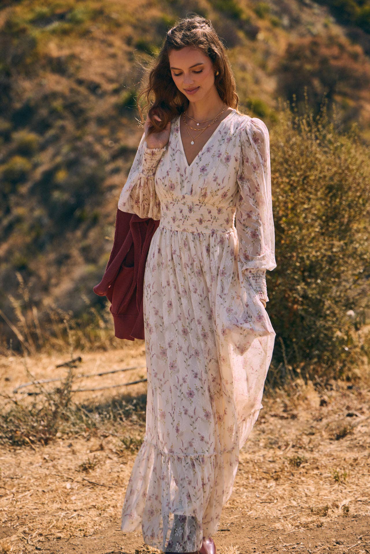 Front view — cream floral chiffon maxi dress with V-neck, sheer long sleeves, smocked waist, ruffle hem; model with mauve sweater over shoulder.
