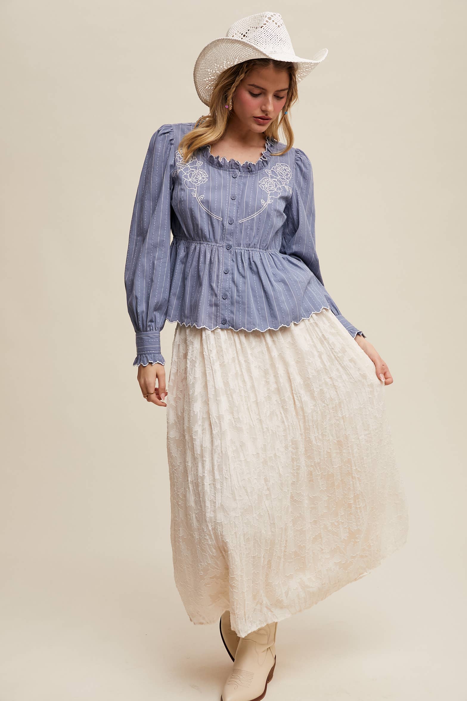 Dusty blue pinstripe blouse with ruffled neckline, white rose embroidery, puff long sleeves, button front, and scalloped peplum hem—styled with a white lace skirt and cowboy hat.