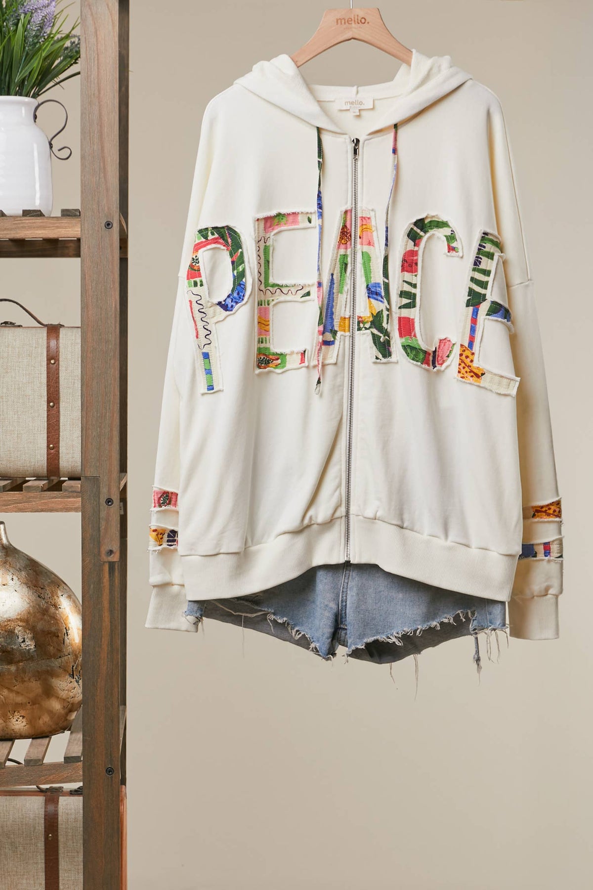 An off-white zip-up hoodie with a colorful 'PEACE' graphic across the front, hanging on a wooden hanger against a beige wall with a rustic wooden shelf on the left.