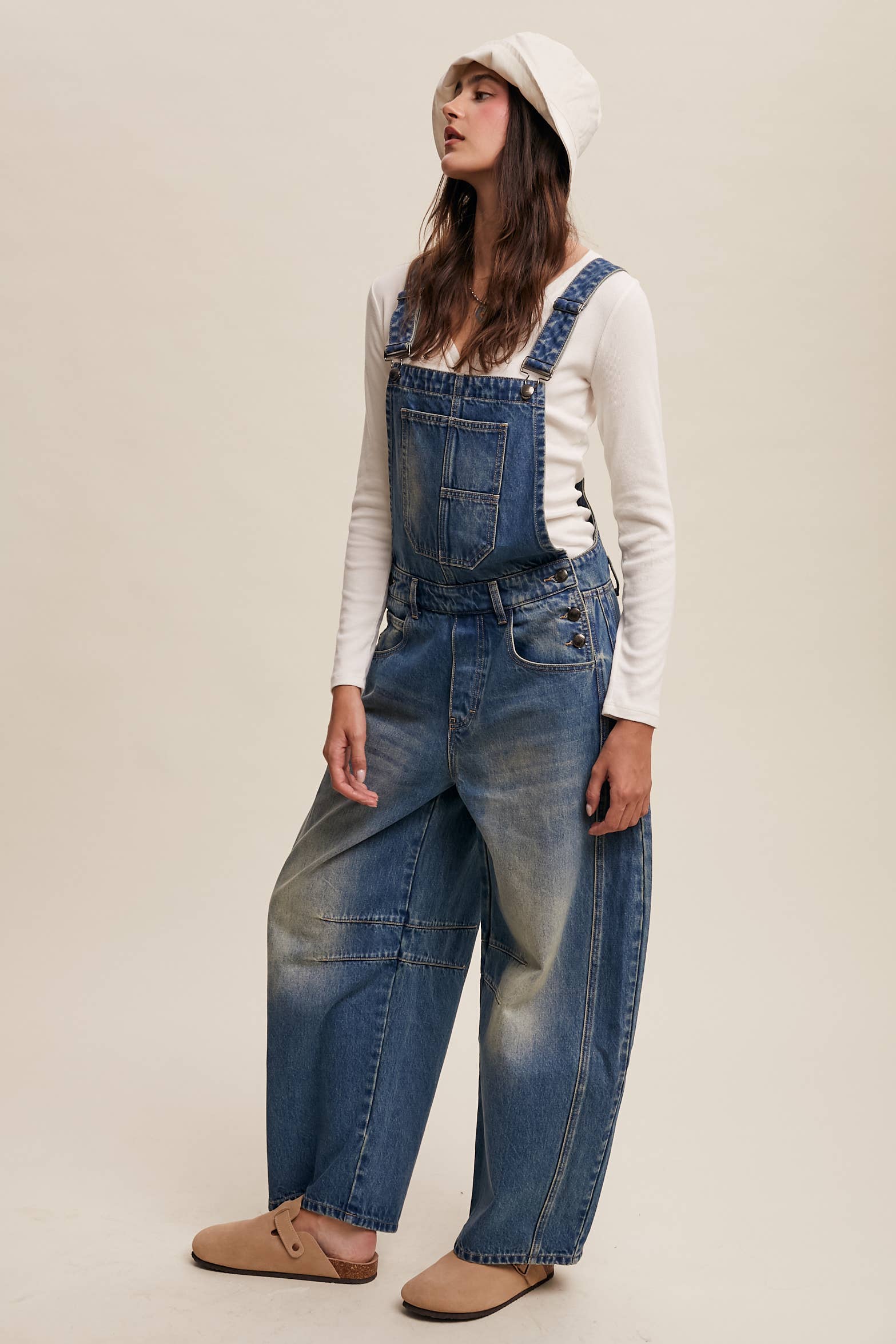 Side view- Vintage-inspired blue denim overalls with relaxed wide-leg fit, front chest pocket, and adjustable straps, styled with a white long-sleeve top and neutral clogs.
