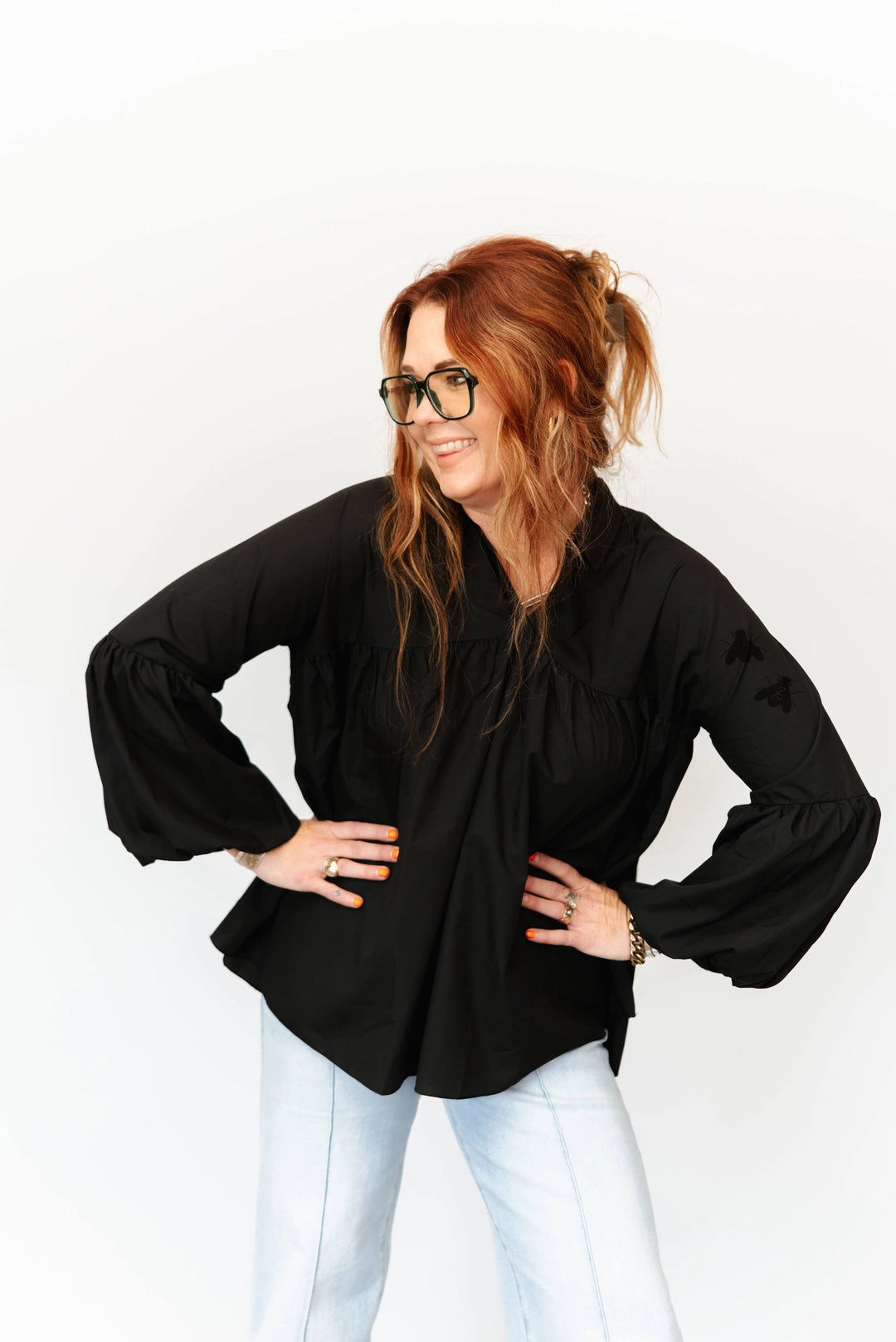 A woman with red hair and glasses wearing a black long-sleeve blouse with balloon sleeves and light blue jeans, posing with hands on hips against a white studio background.