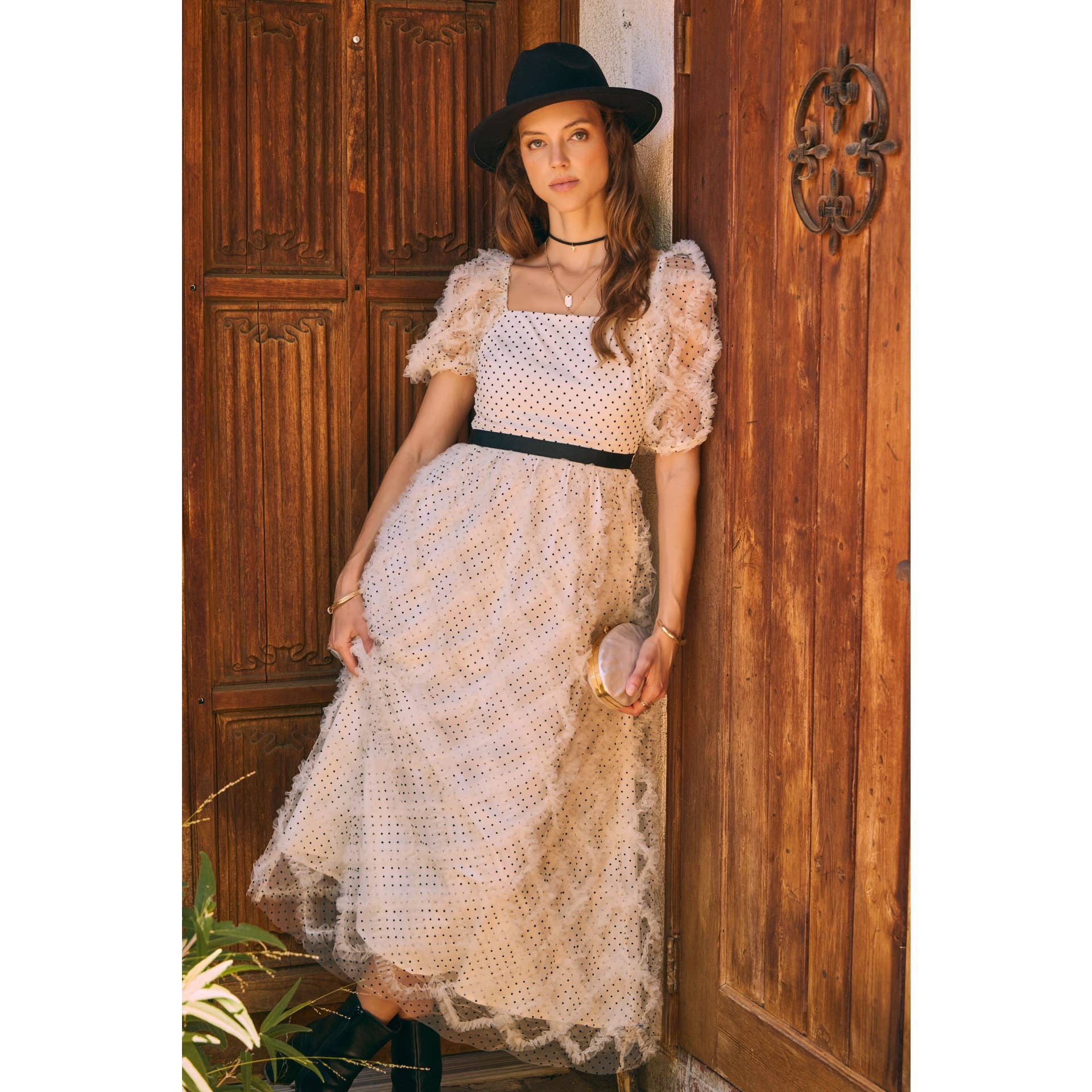 Front view — cream polka-dot tulle midi dress with puff sleeves and black belt; styled with black hat, ankle boots, pearl clutch; leaning by wooden doors.