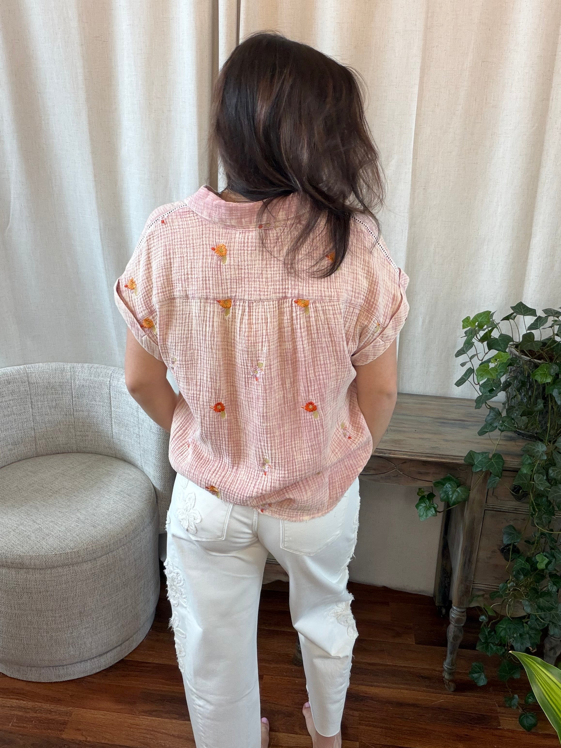 Person wearing a pink blouse with orange buttons and white pants in a room with a curtain and plant.