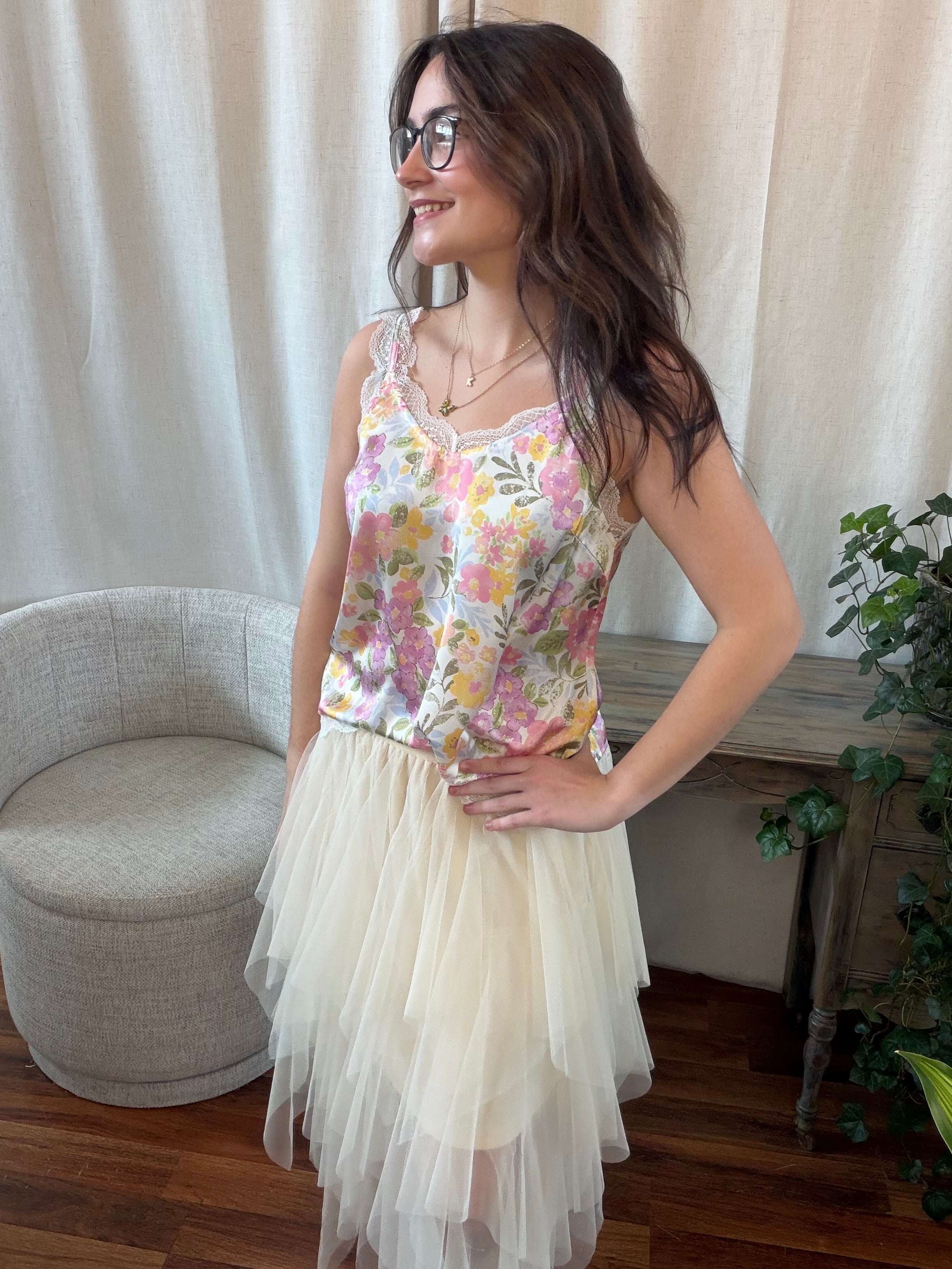 Woman wearing a floral top and white skirt standing in a room with a chair and plants.