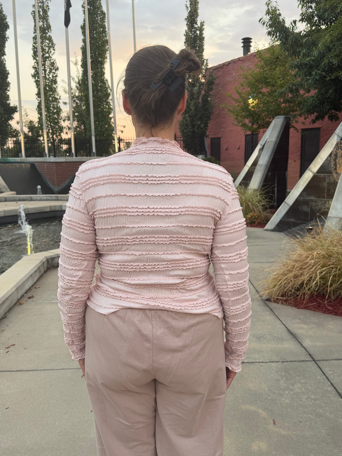 A woman viewed from behind wearing a light pink textured long-sleeve top and beige pants, standing outdoors on a concrete walkway near a building and trees.
