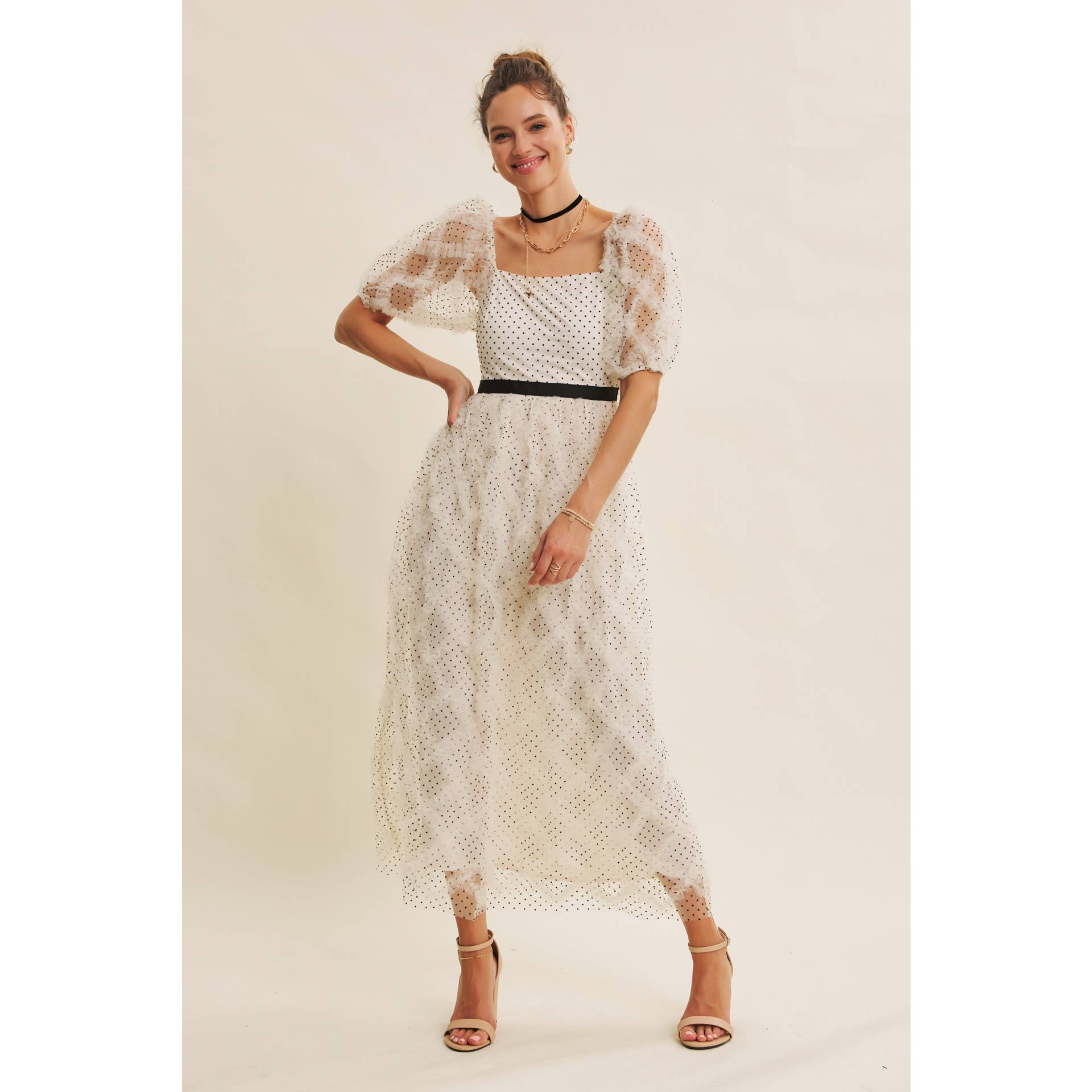 Front view — cream polka-dot tulle midi dress with puff sleeves and black belt; styled with black hat, ankle boots, pearl clutch; leaning by wooden doors.