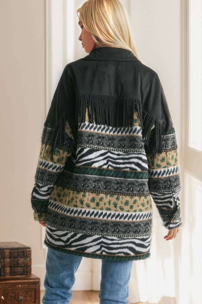 Brown Aztec print shacket with fringe detail, button closure, and patterned sleeves, styled with cream pleated pants while seated on a sofa.