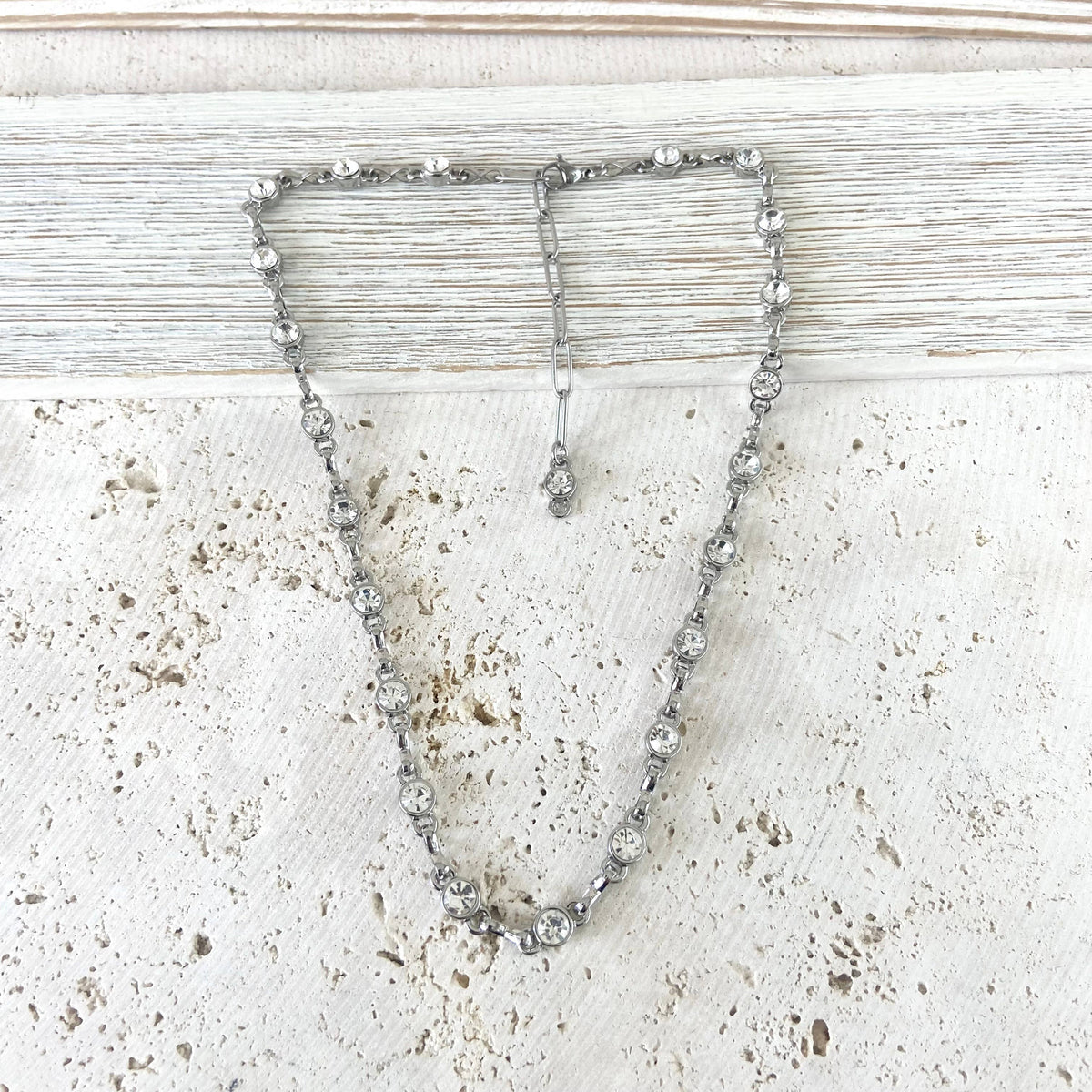 A silver-toned beaded necklace with clear faceted rhinestones, draped in a V-shape on a distressed white wooden surface.