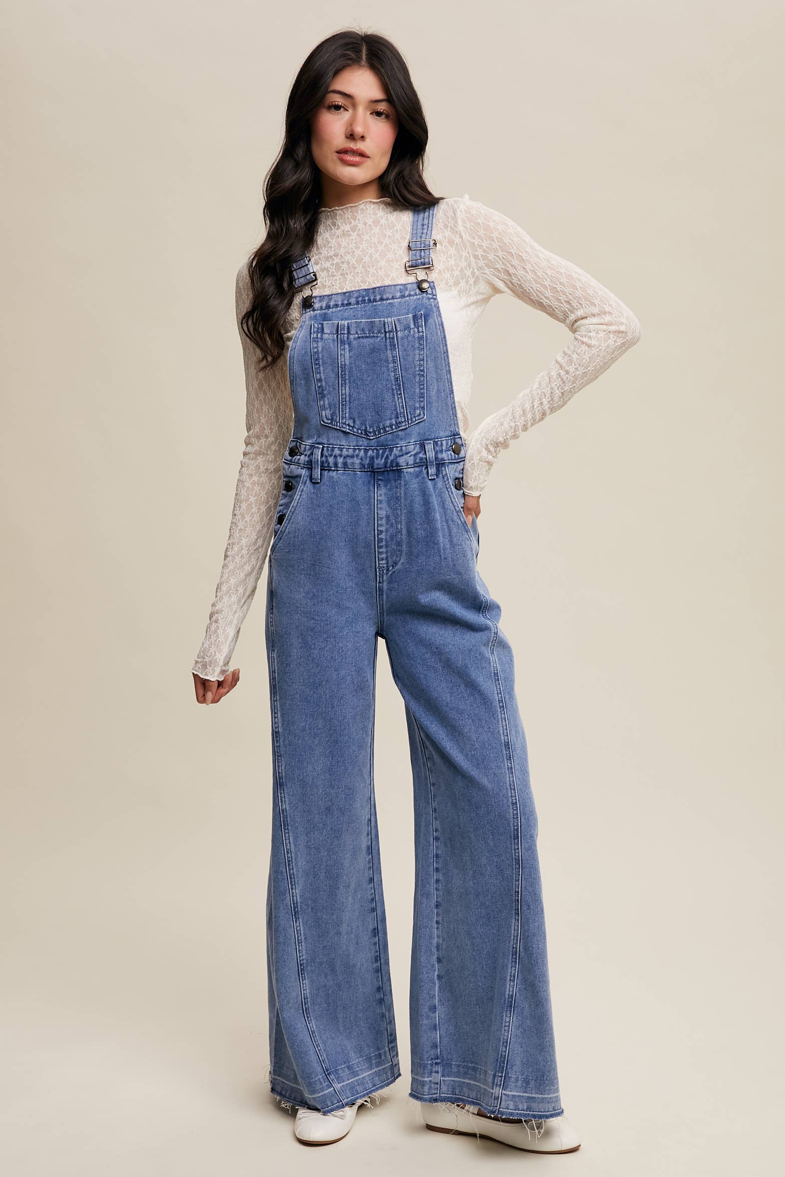 Light-wash wide-leg denim overalls with adjustable straps, front bib pocket, side buttons, and slant pockets, finished with a raw hem and layered over a cream long-sleeve top.