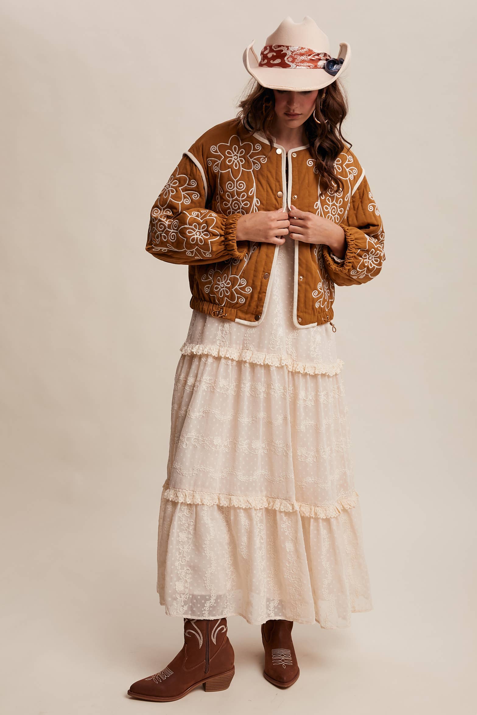 Boho caramel-brown jacket with cream floral swirl embroidery, ivory piping, and snap buttons, styled over a lace tie-front top with a scarf and felt hat outdoors.