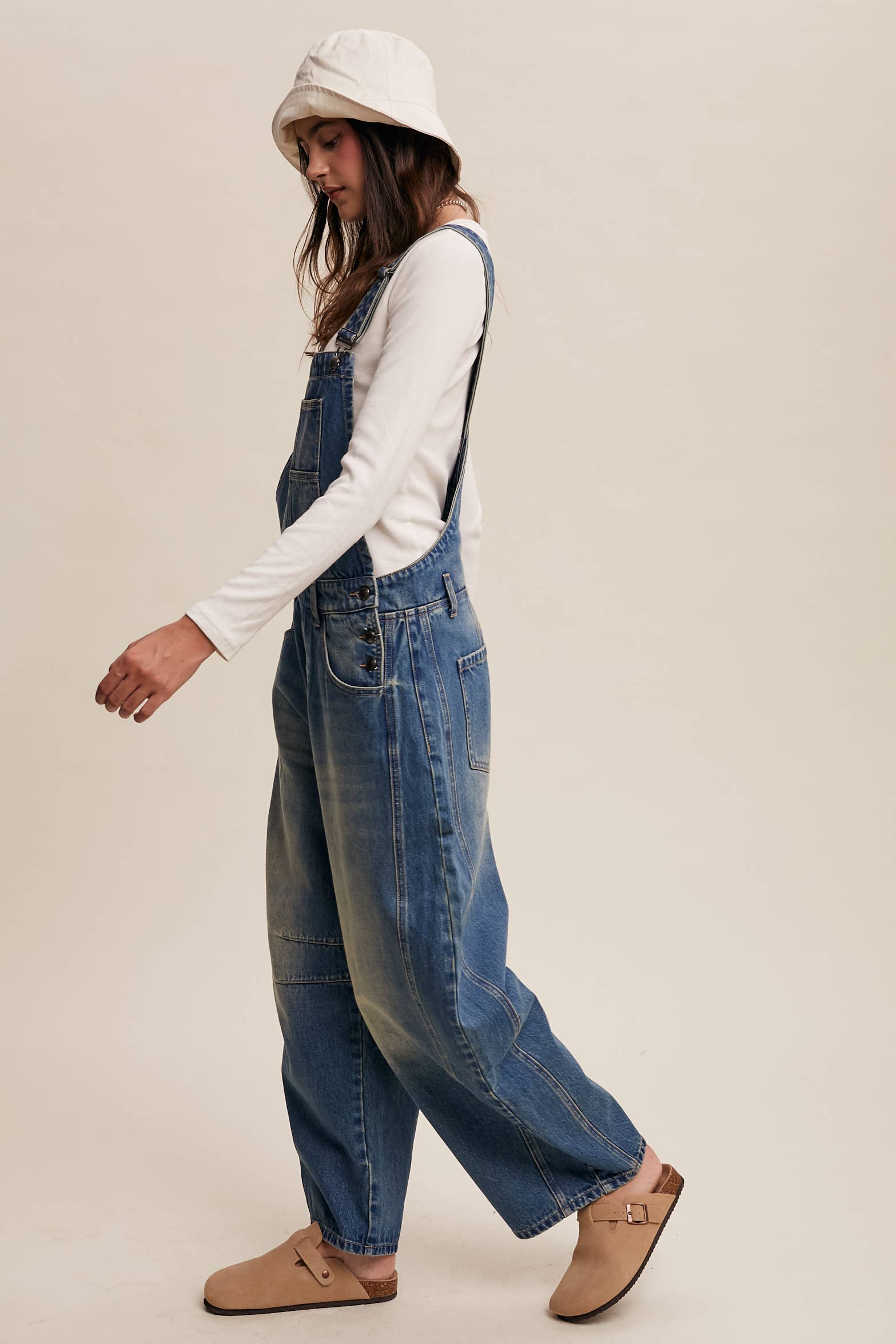 Side view- Vintage-inspired blue denim overalls with relaxed wide-leg fit, front chest pocket, and adjustable straps, styled with a white long-sleeve top and neutral clogs.
