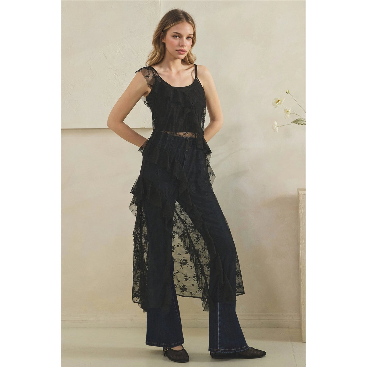 A woman modeling a black sheer lace duster over dark wide-leg jeans, standing against a light beige wall.