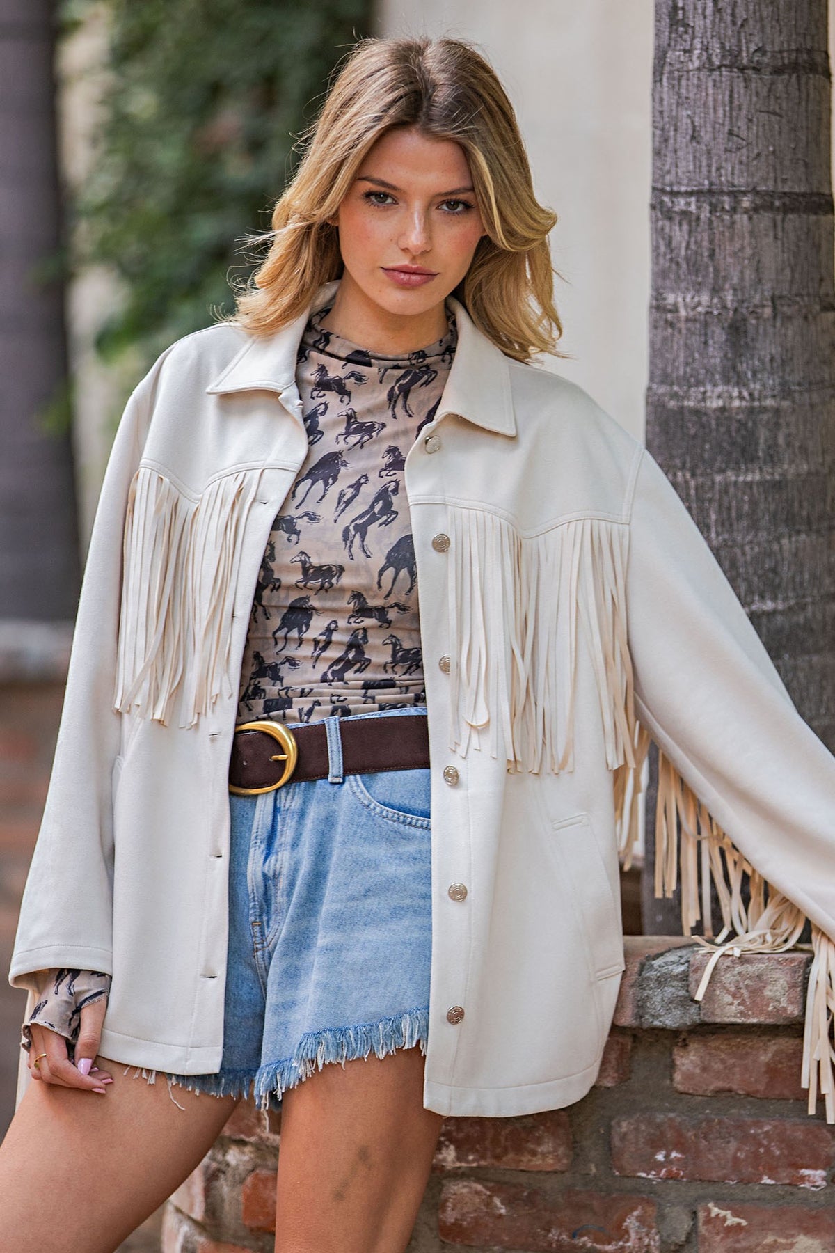A woman models a cream-colored faux leather fringe jacket with a button-down front, worn over a patterned t-shirt and denim shorts.