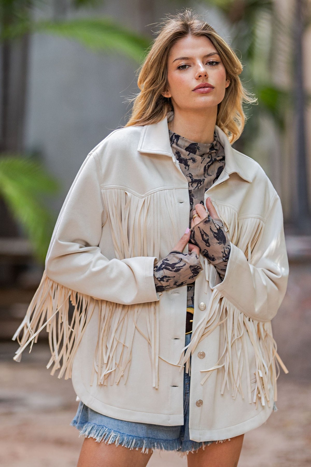 A woman wearing a cream-colored fringe jacket over a patterned top and denim shorts, standing outdoors with a blurred background.
