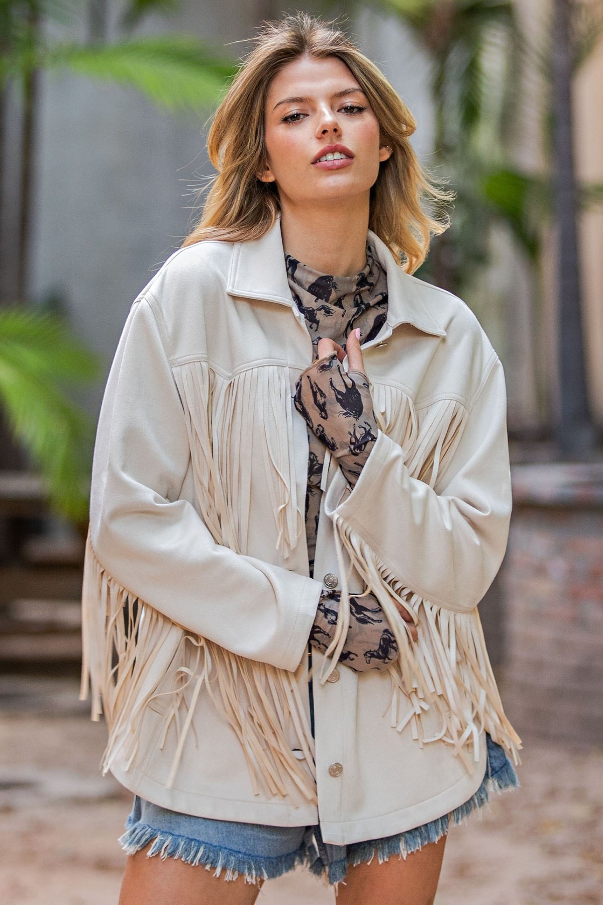 A woman wearing a cream-colored faux leather fringe jacket over a patterned top and denim shorts, standing outdoors with a blurred background of palm trees and brick.