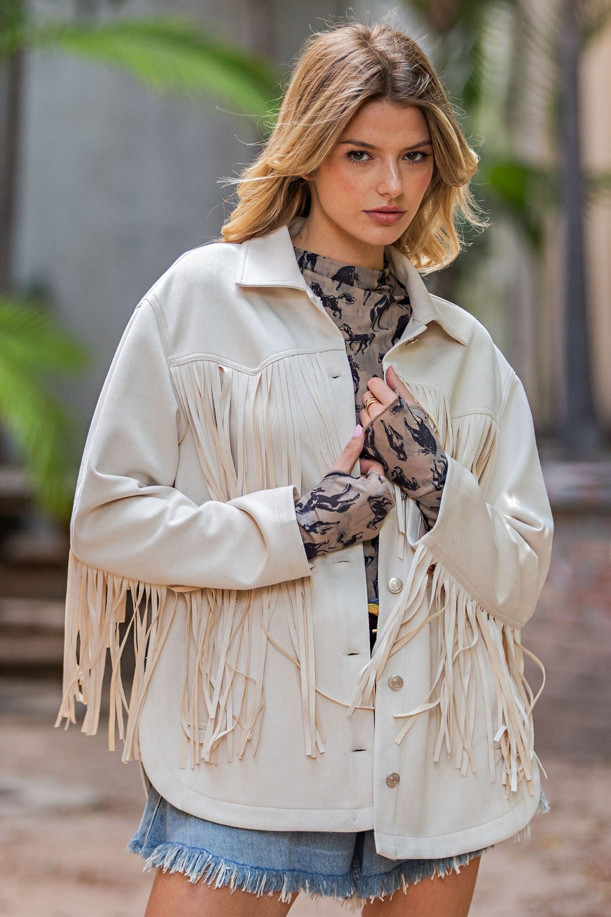 A woman wearing a cream-colored faux leather fringe jacket over a patterned top and denim shorts, standing outdoors with a blurred background.