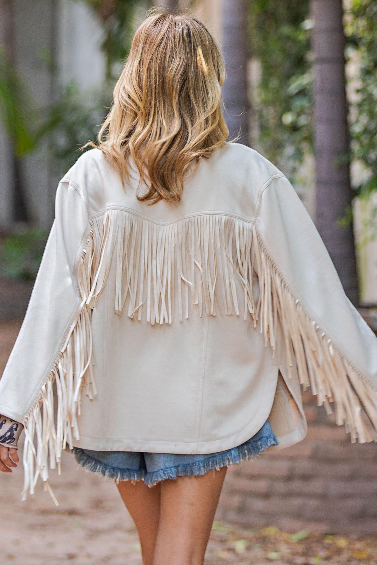 A woman viewed from behind wearing a cream-colored fringe jacket over denim shorts, walking outdoors in a park-like setting with blurred trees in the background.