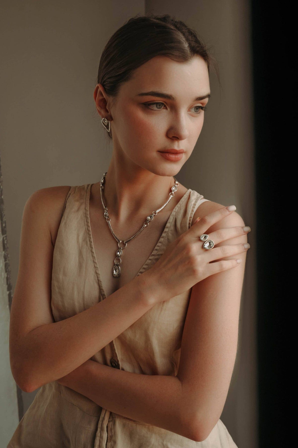 A woman wearing a silver-toned pendant necklace and matching ring, posed in a warm beige dress against a soft neutral background.