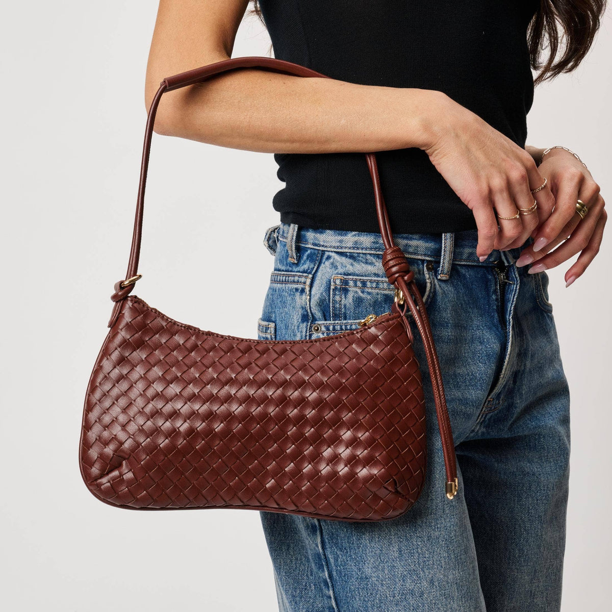 Cindy Woven Crescent Shoulder Bag in Burgundy by Moda Luxe — close-up model carry view showing woven vegan leather crescent silhouette, knotted strap detail, and gold-tone zip pull, styled with black top and denim jeans