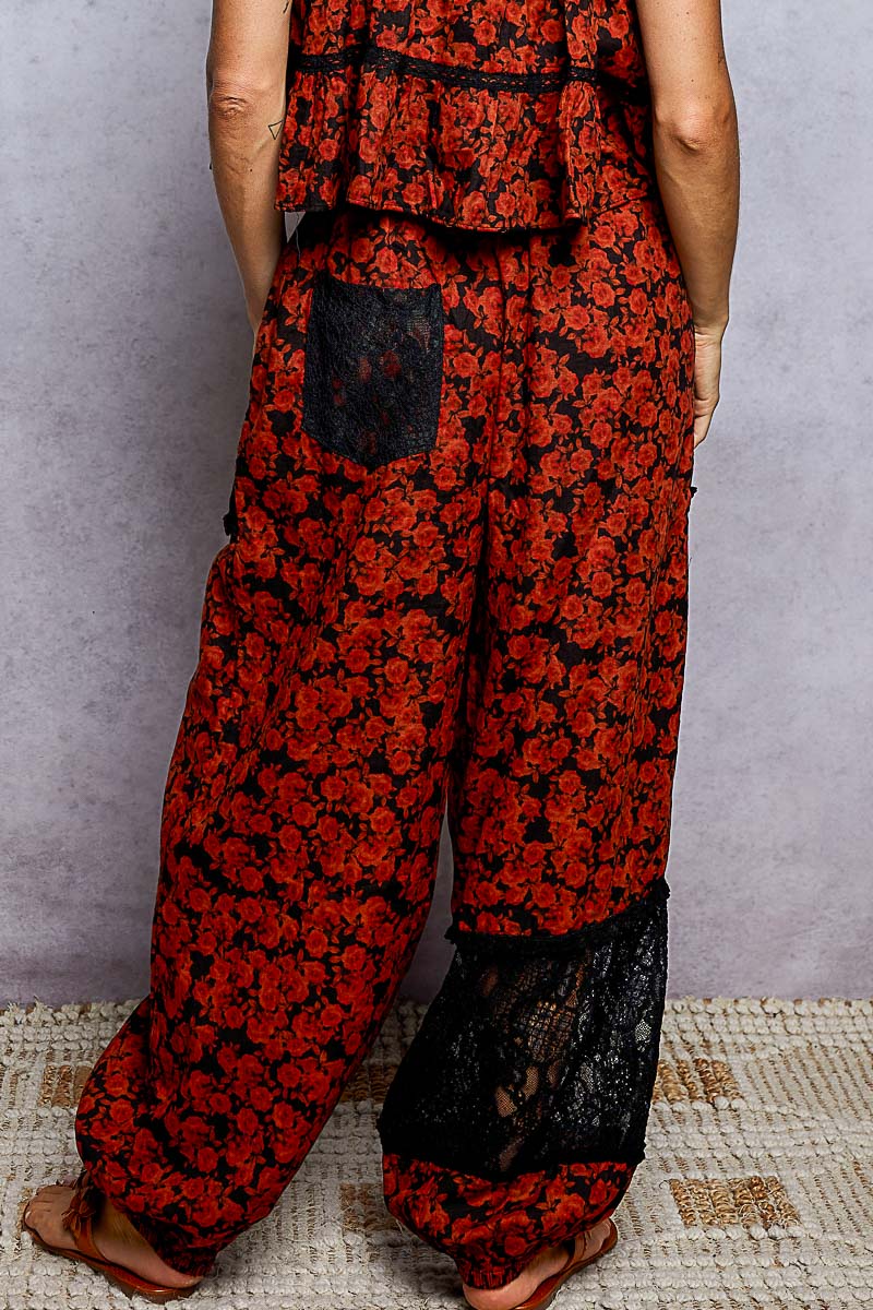 A person wearing loose, wide-leg pants with a red and black floral print and black lace panel details at the back and lower hems, shown from the back against a gray wall.