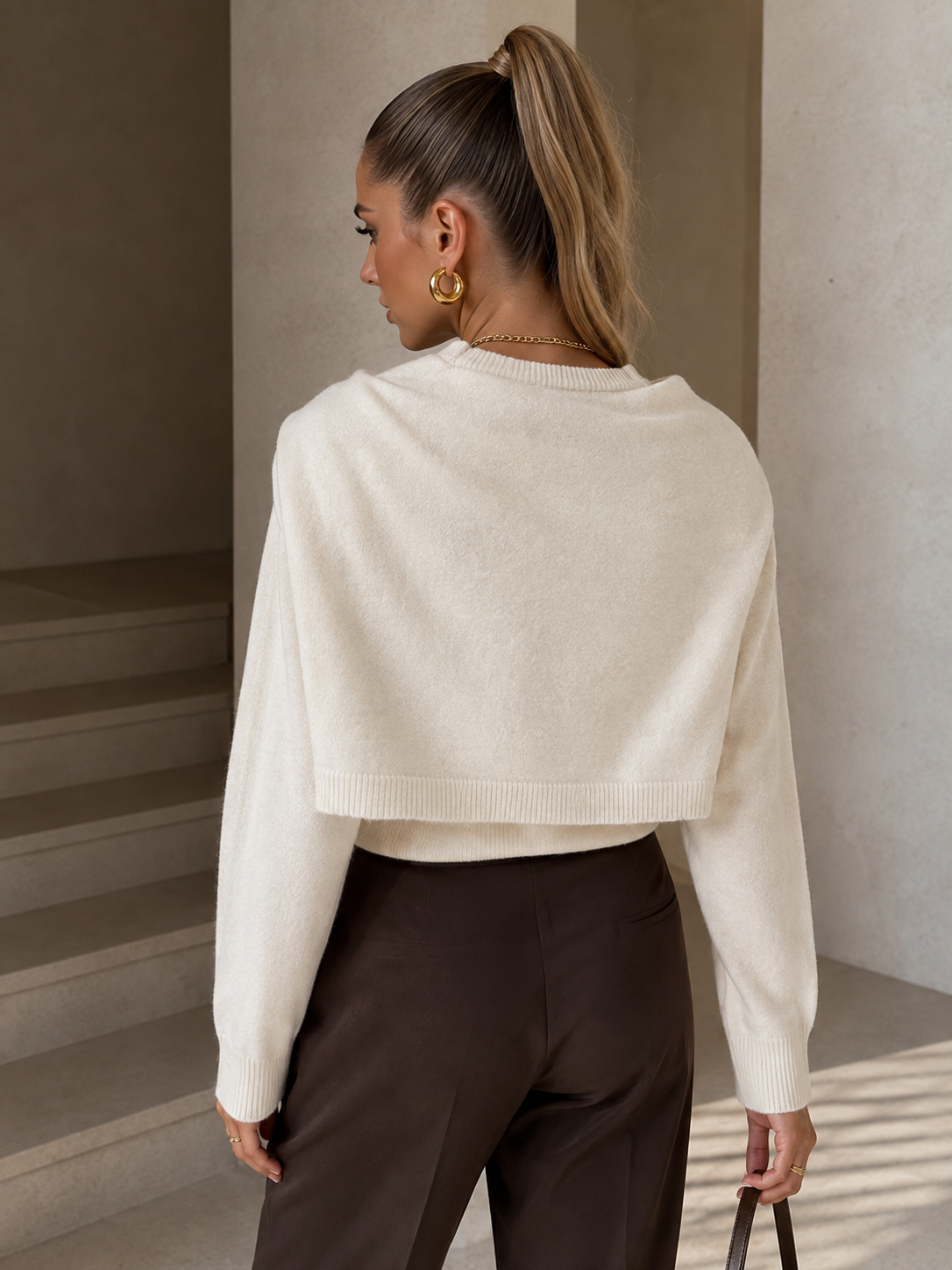 A woman shown from behind wearing a cream-colored cropped knit sweater and dark brown trousers, standing in a minimalist indoor setting with concrete walls and stairs.