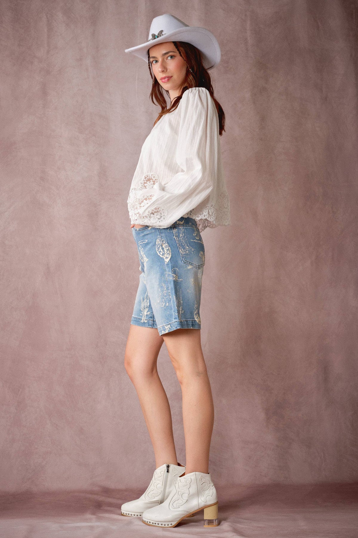 A woman modeling a white long-sleeve blouse with lace trim, light-wash distressed denim shorts, and white heeled boots, standing against a textured dusty pink studio backdrop.