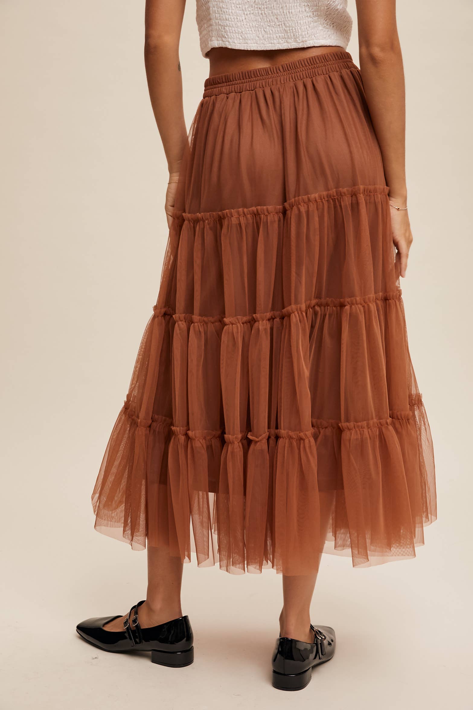 Back view — rust tulle tiered midi skirt with elastic waist and ruffle tiers, sheer overlay; model in black Mary Jane flats.