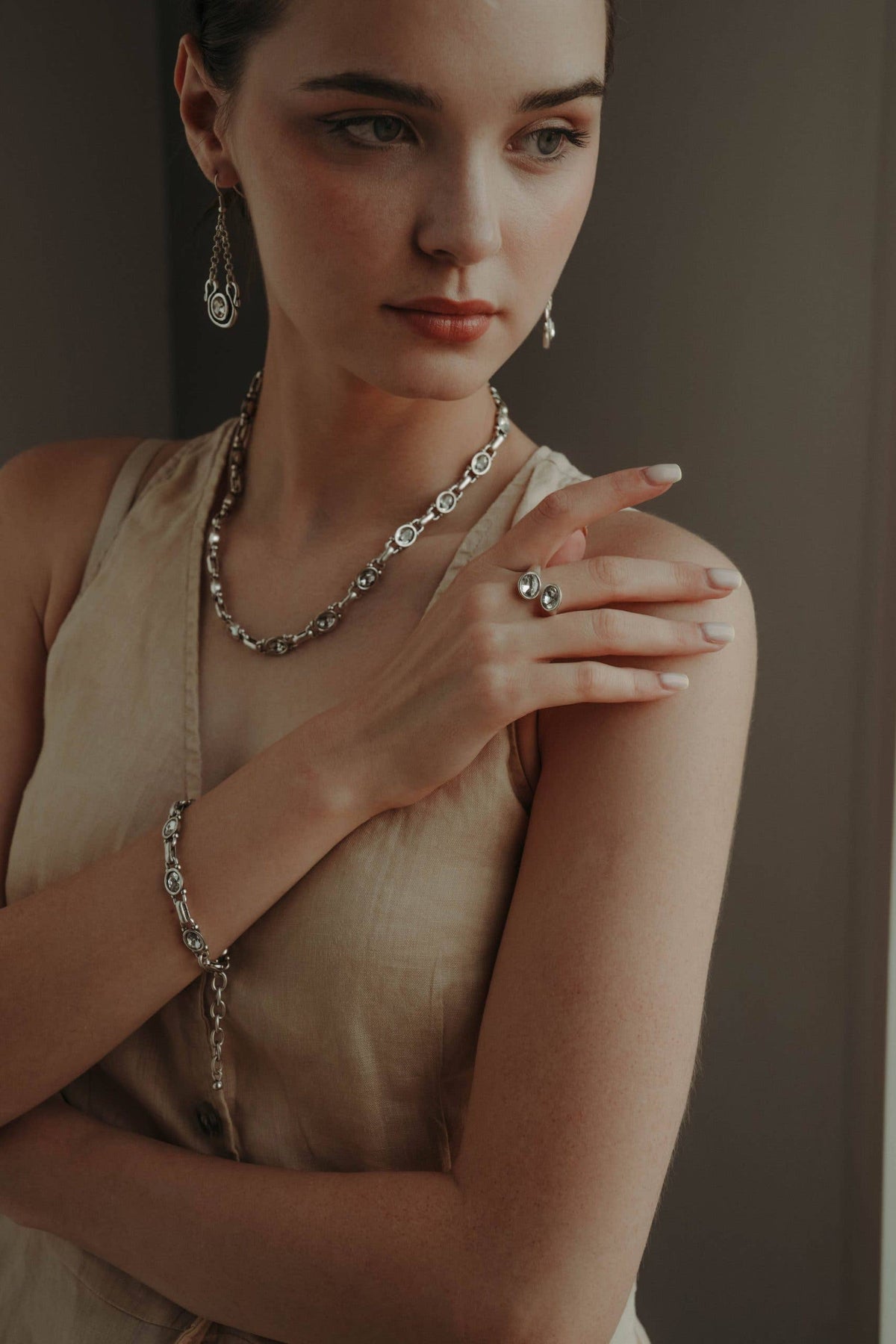 A woman wearing a matching silver-toned jewelry set including a chain necklace, bracelet, and dangling earrings, styled with a silver ring on her finger. She is wearing a beige sleeveless top against a dark gray background.