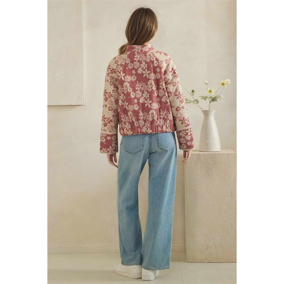 A woman stands with her back to the camera wearing a dusty rose and cream floral patterned jacket and light blue wide-leg jeans, standing in a minimalist beige room next to a white vase with flowers.
