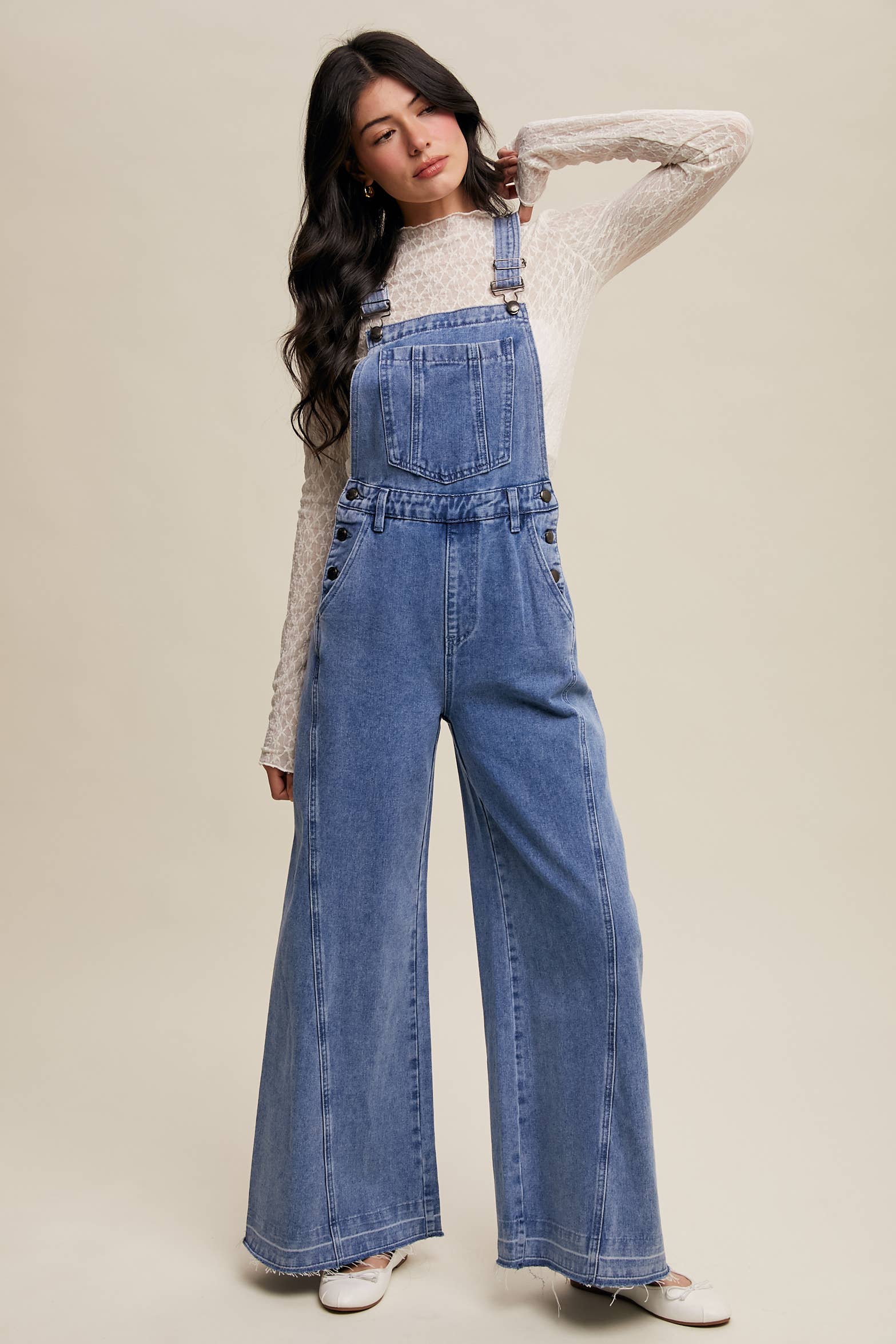 Light-wash wide-leg denim overalls with adjustable straps, front bib pocket, side buttons, and slant pockets, finished with a raw hem and layered over a cream long-sleeve top.