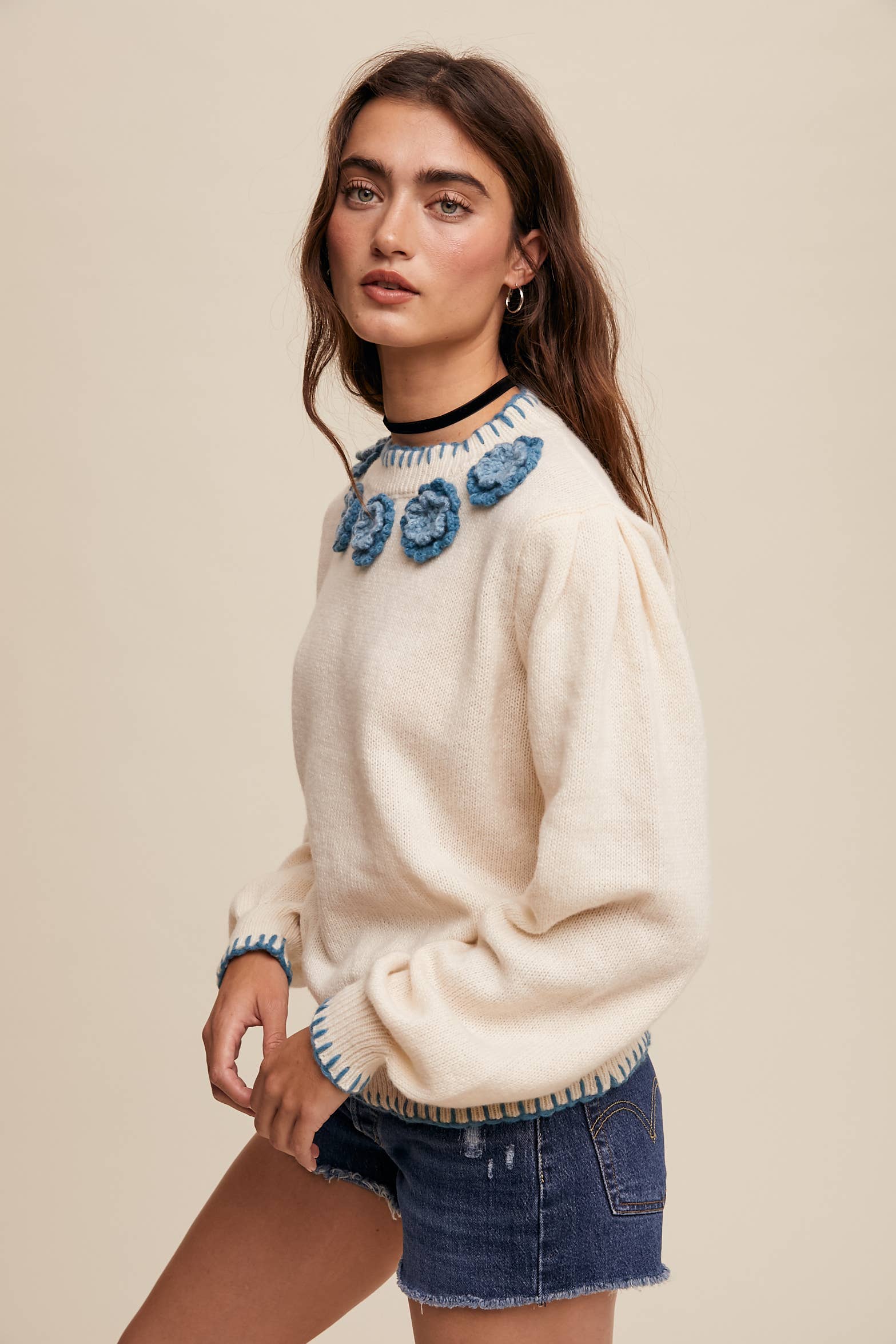 Side view- Cream sweater with blue crochet flower appliqués at neckline, blue whipstitch trim at cuffs and hem, long sleeves; styled with frayed denim shorts.
