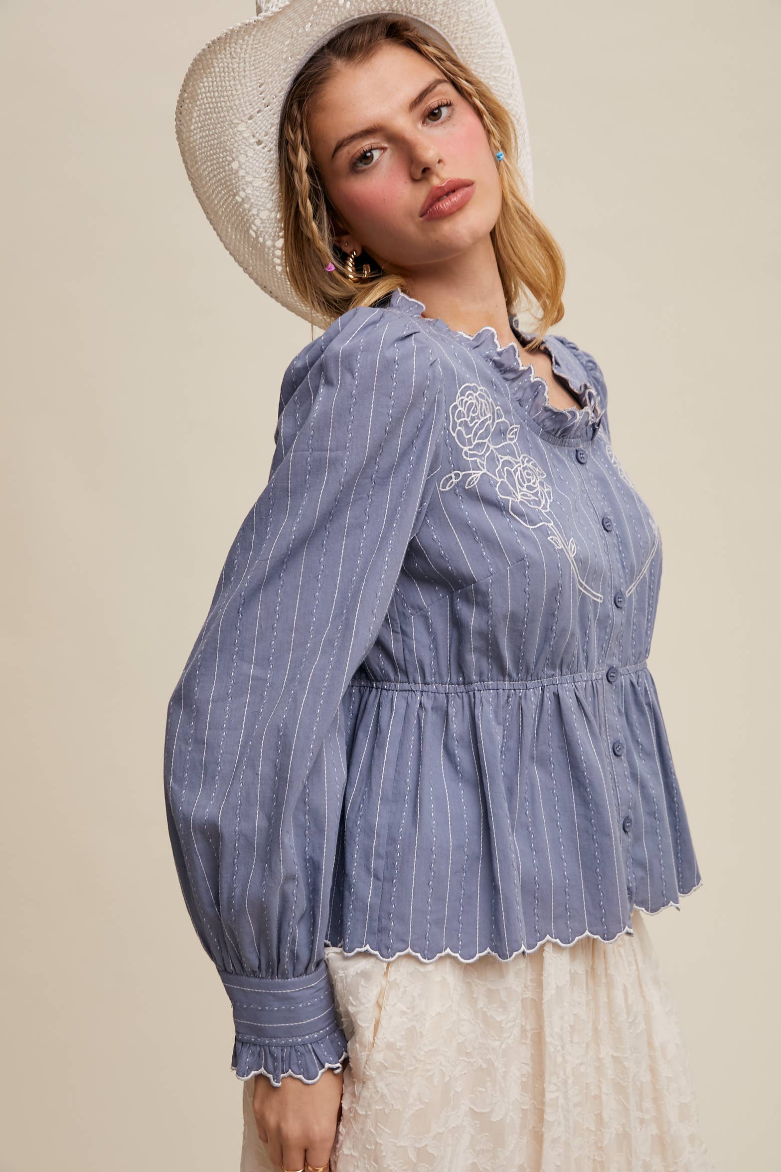 Side view- Dusty blue pinstripe blouse with ruffled neckline, white rose embroidery, puff long sleeves, button front, and scalloped peplum hem—styled with a white lace skirt and cowboy hat.