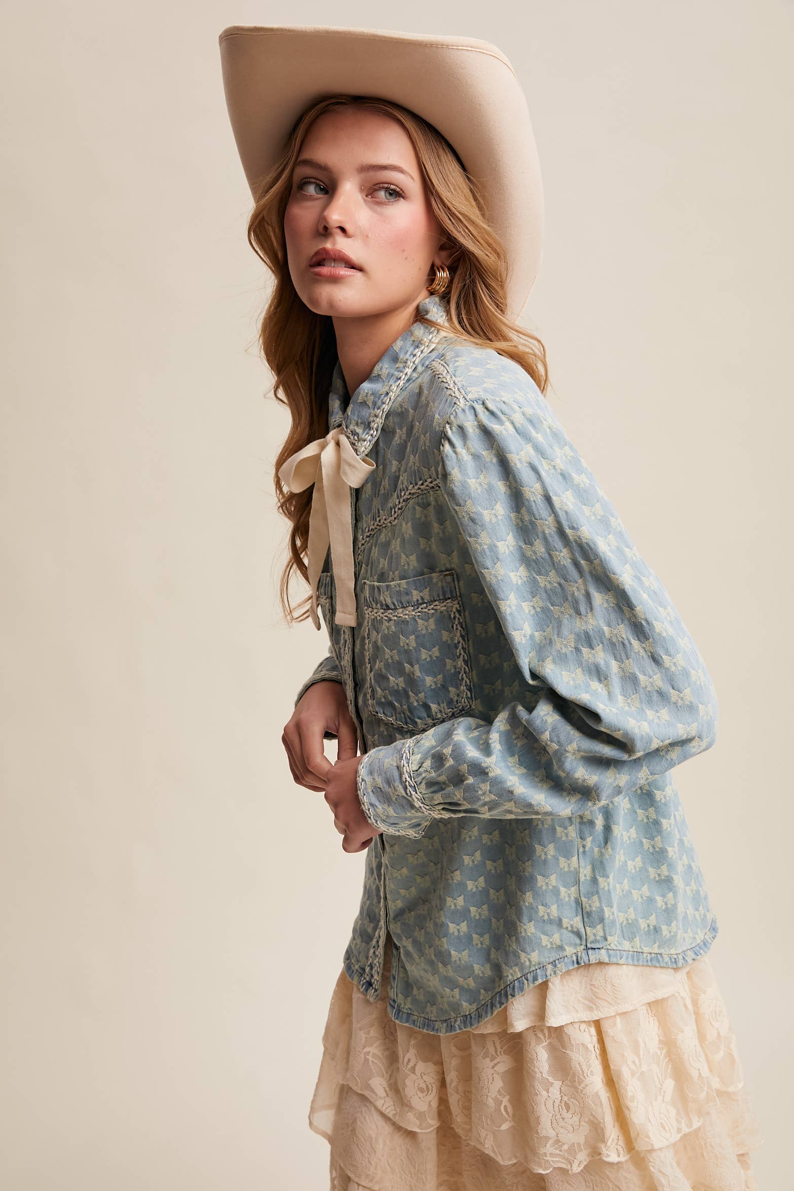 Side view- Light-wash denim blouse with cream bow-tie collar, tonal mini-bow pattern, braided contrast stitching, snap front, and two chest pockets.