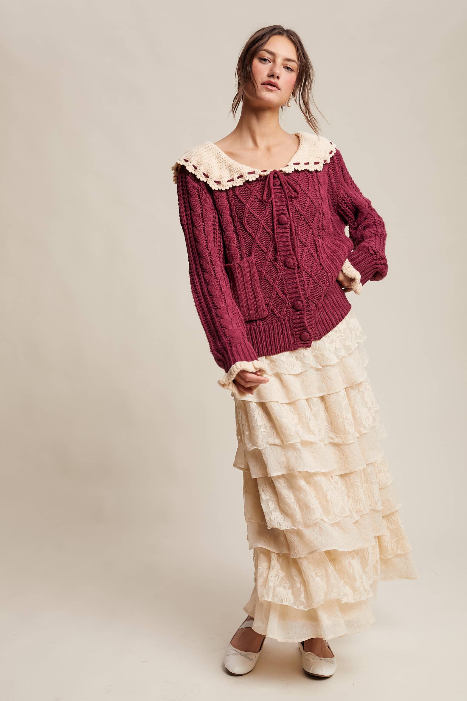 Woman wearing a burgundy cable knit sweater and beige layered skirt on a plain background