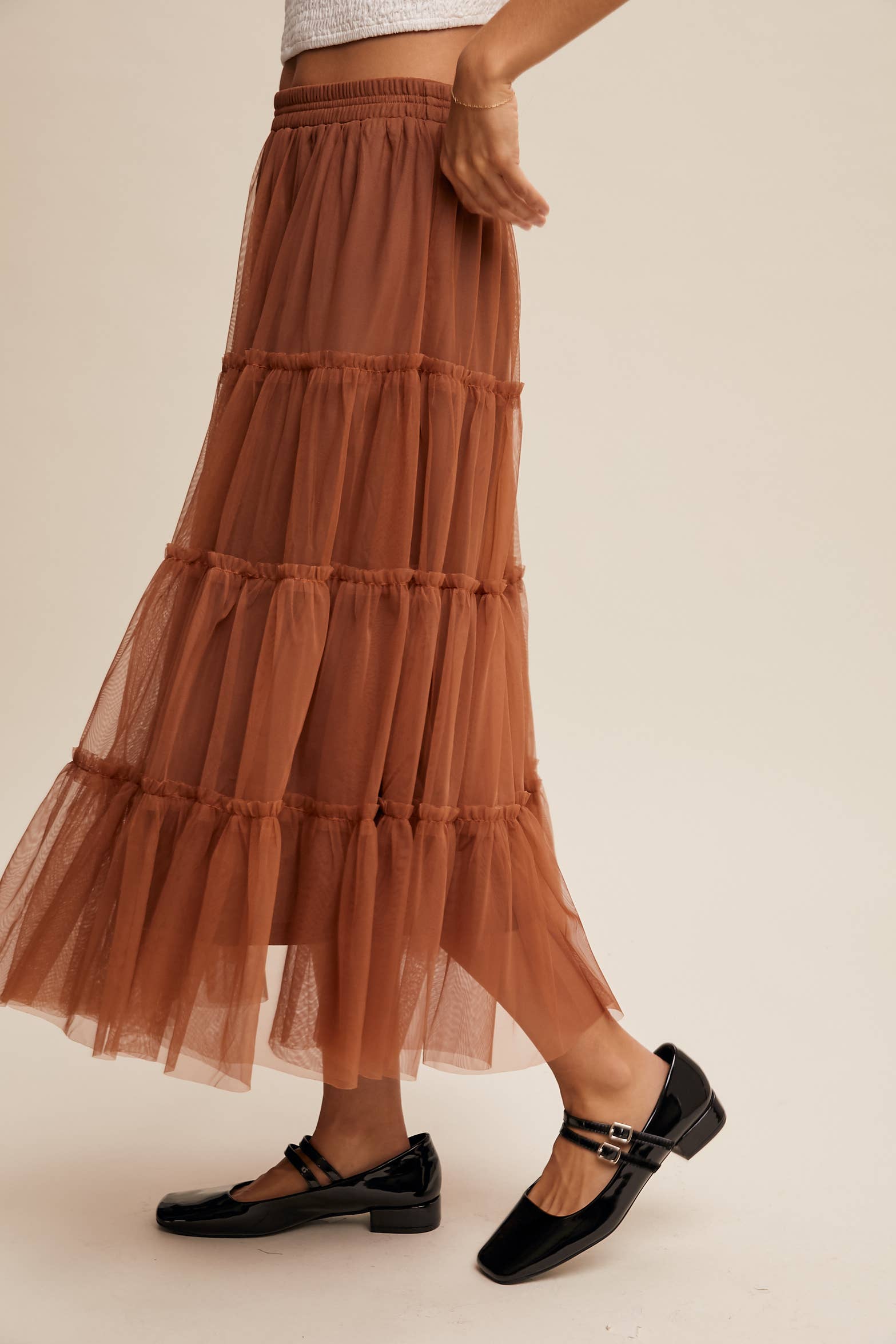 Side view — rust tulle tiered midi skirt with elastic waist and ruffle tiers, sheer overlay; model in black Mary Jane flats.