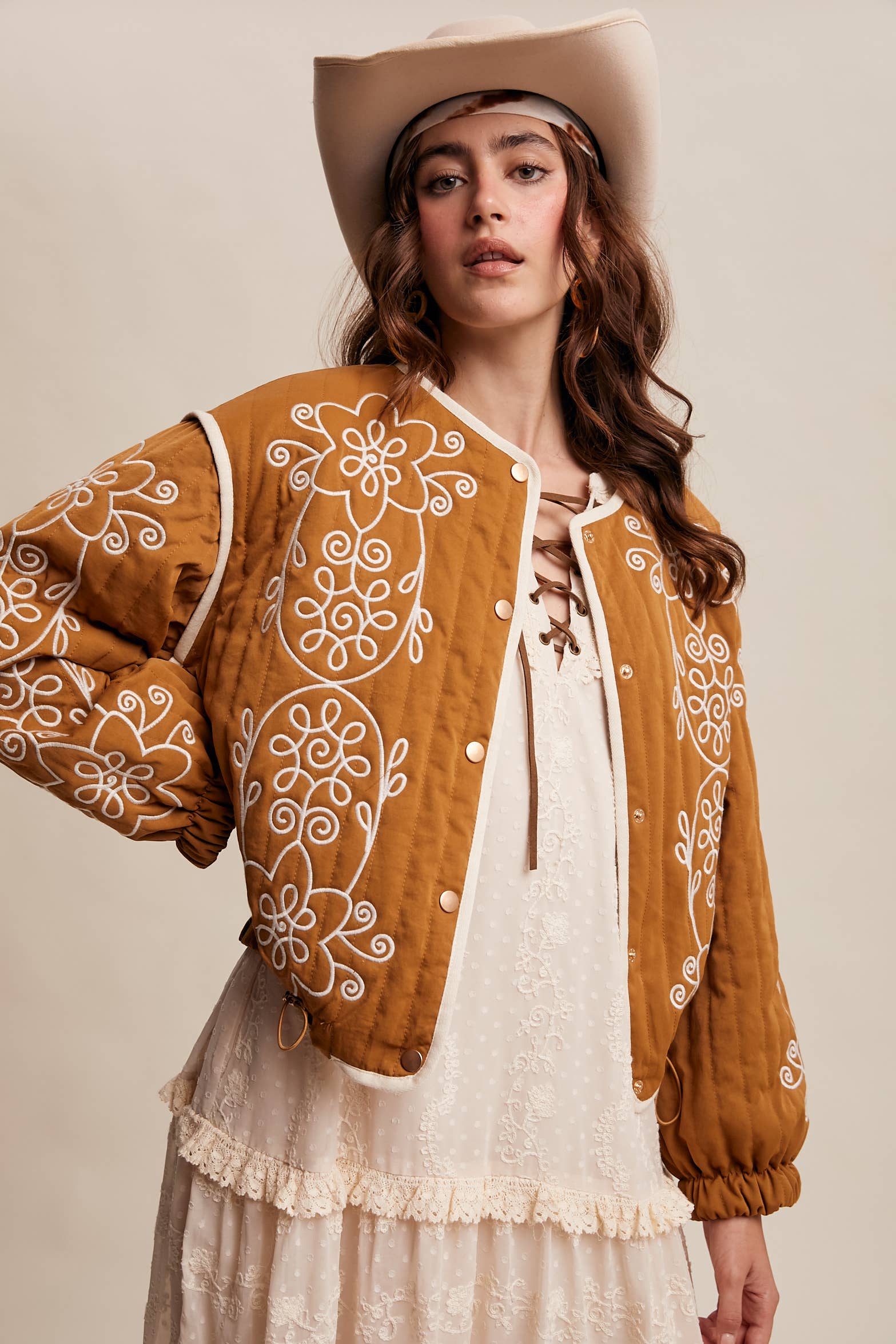 Boho caramel-brown jacket with cream floral swirl embroidery, ivory piping, and snap buttons, styled over a lace tie-front top with a scarf and felt hat outdoors.