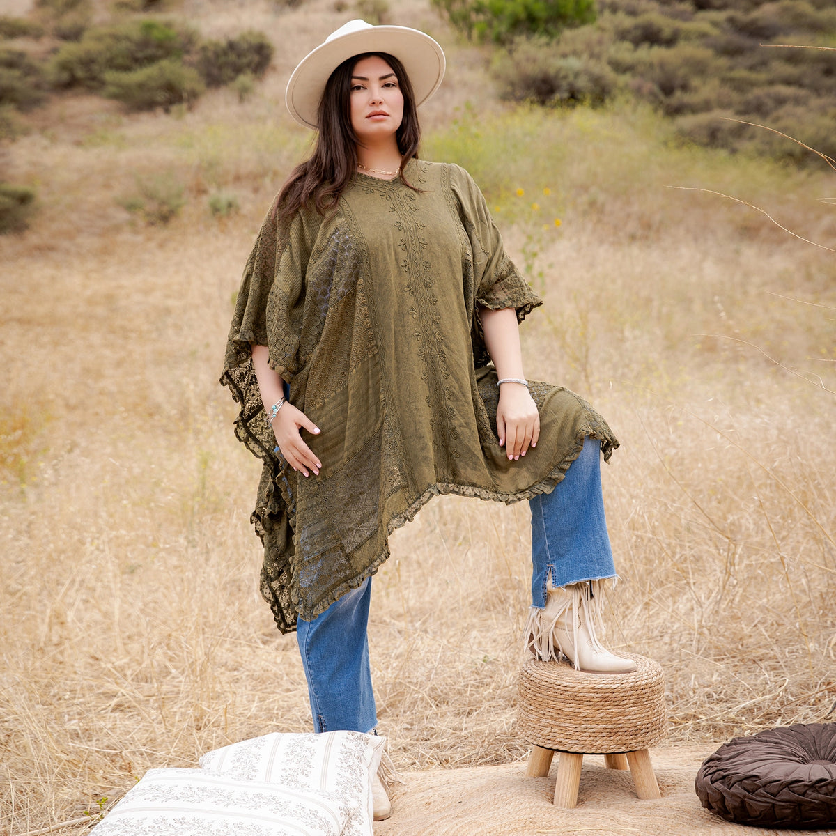 A woman stands outdoors in a dry grassy field wearing a loose olive green bohemian tunic top, blue jeans, white ankle boots, and a wide-brimmed cream hat, with a small woven stool beside her.