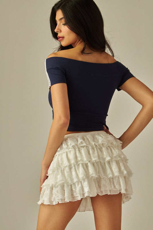Back view of women’s white lace tiered ruffle mini skirt with flouncy layers, styled with black ankle-strap heels.
