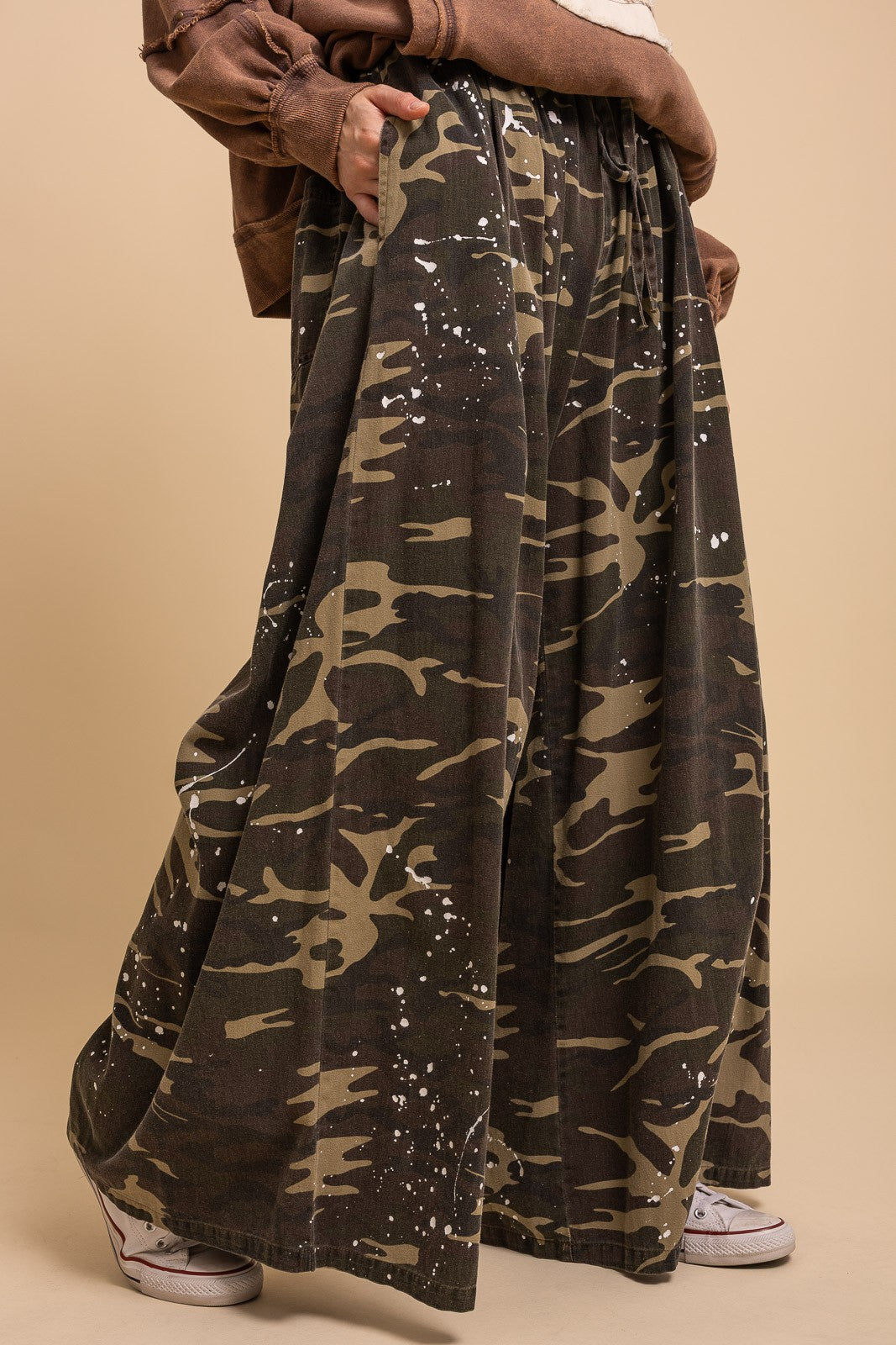 Side close-up of Blue Velvet paint splash camo wide leg pants showing relaxed wide leg silhouette and paint splash camo print, brown jacket and white sneakers