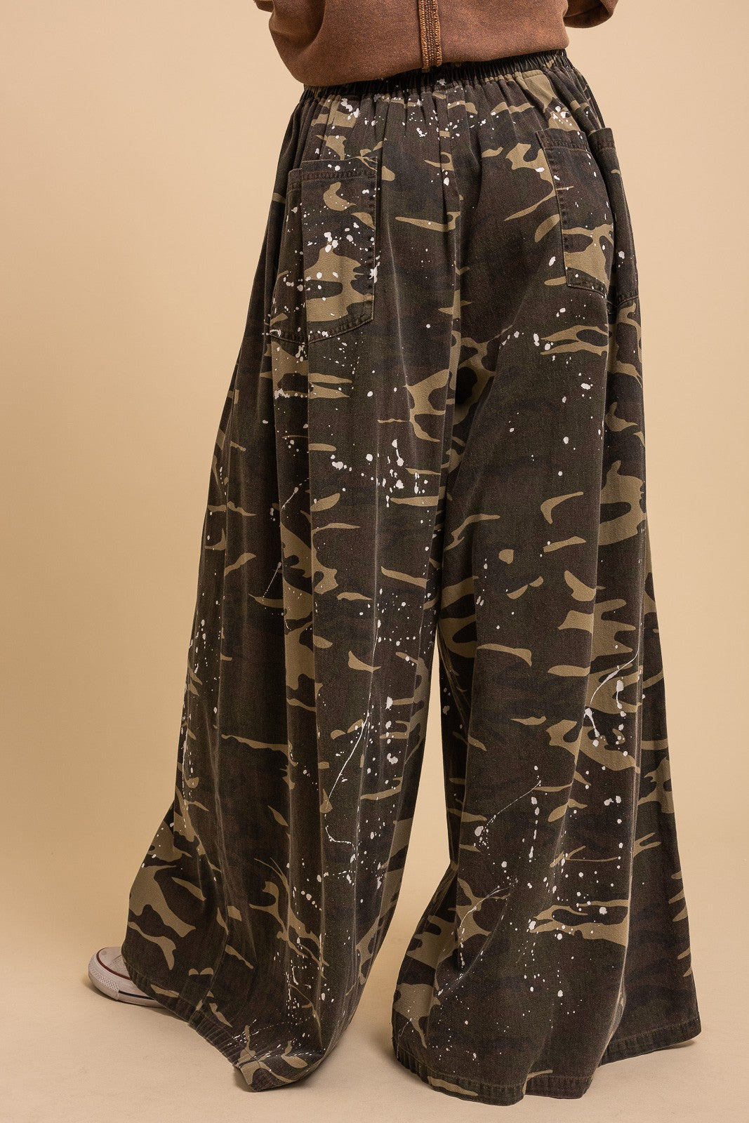 Back view of Blue Velvet paint splash camo wide leg pants showing back pocket detail, elastic waistband, and white paint splash print, styled with brown jacket