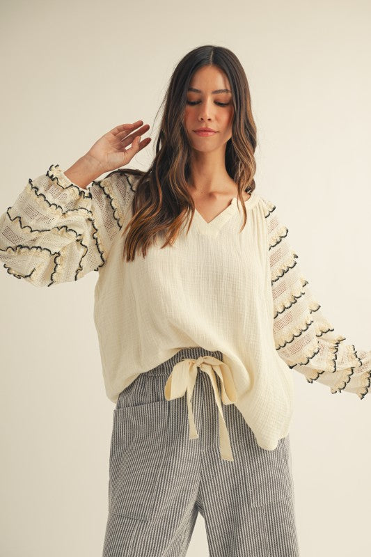Front view of cream gauze blouse with sheer lace balloon sleeves, black-trimmed scalloped ruffles and a V-neck, styled with gray striped wide-leg pants.